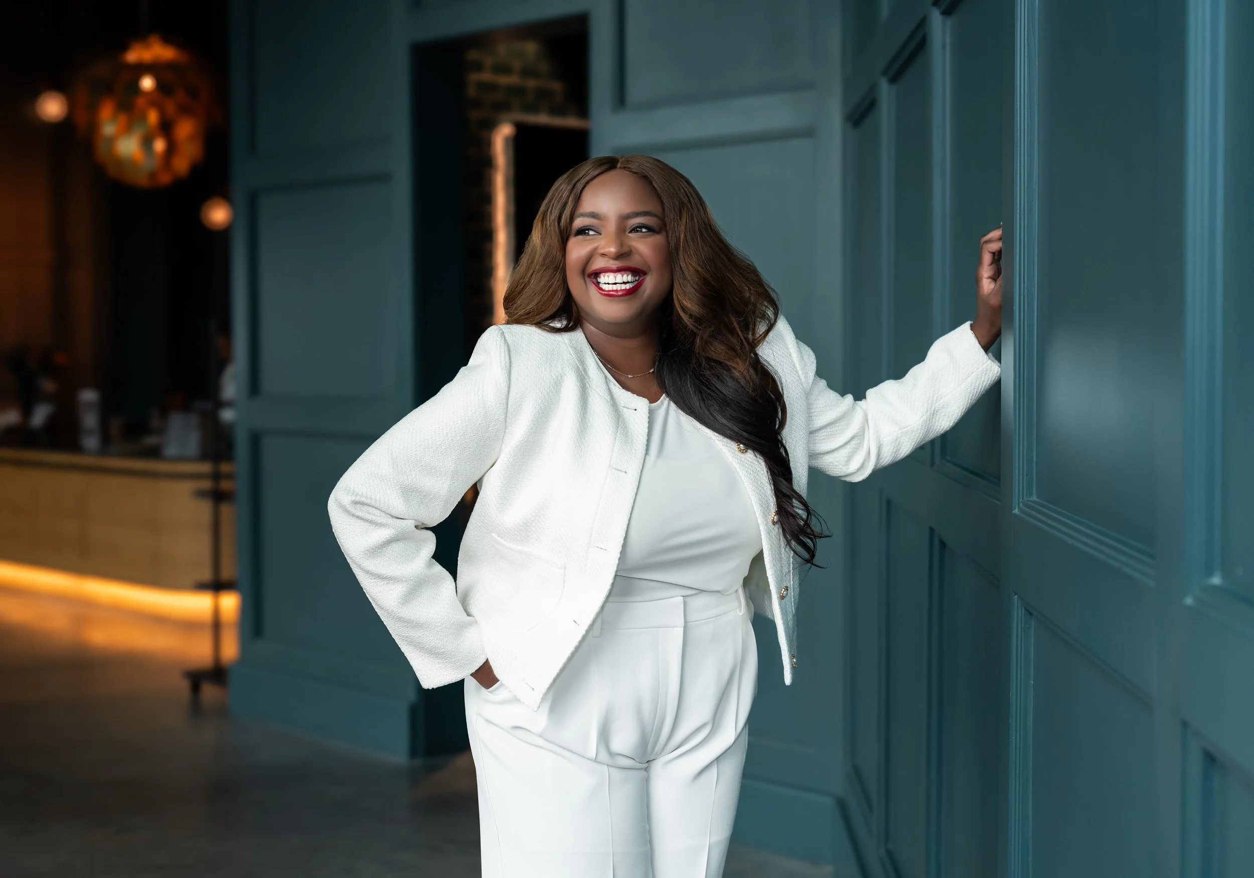 A woman with long, wavy hair, wearing a white blazer and matching pants, smiling and standing in a stylish, modern interior with teal paneled walls.