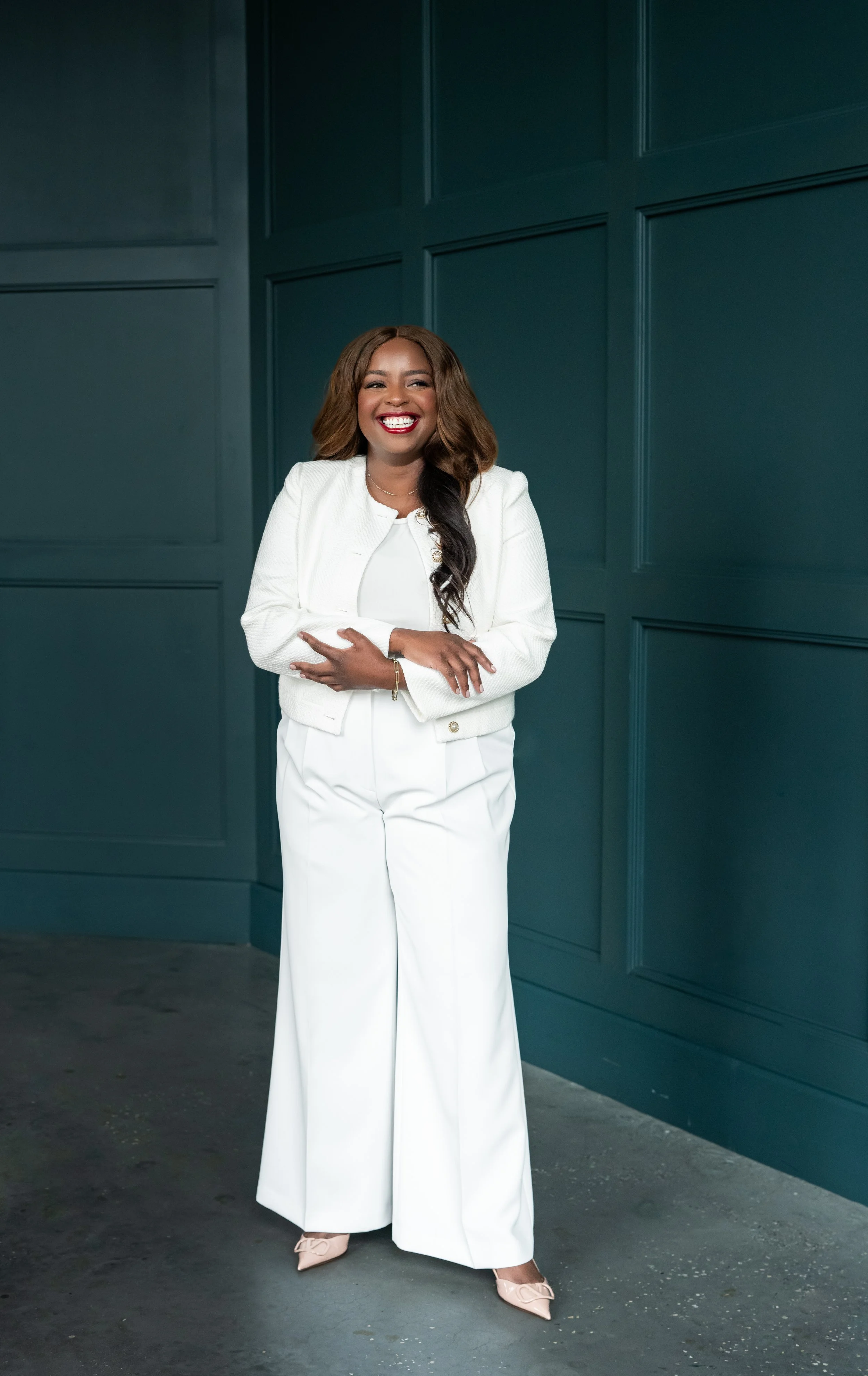 A woman smiling and standing with arms crossed against a teal wall, wearing a white jacket, white wide-leg pants, and beige heels.