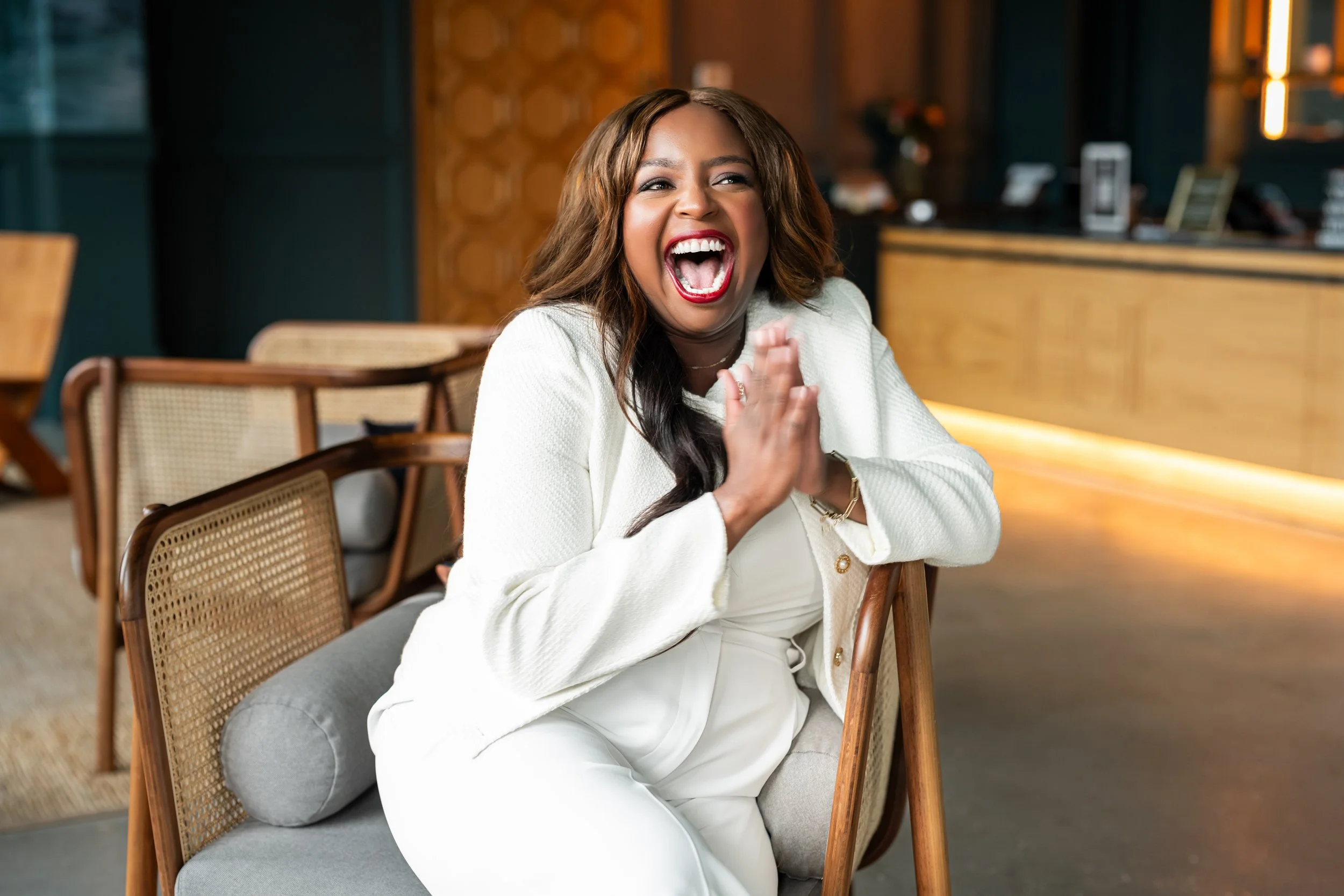 Woman laughing happily, sitting in a modern indoor space, wearing a white outfit with wooden furniture around.