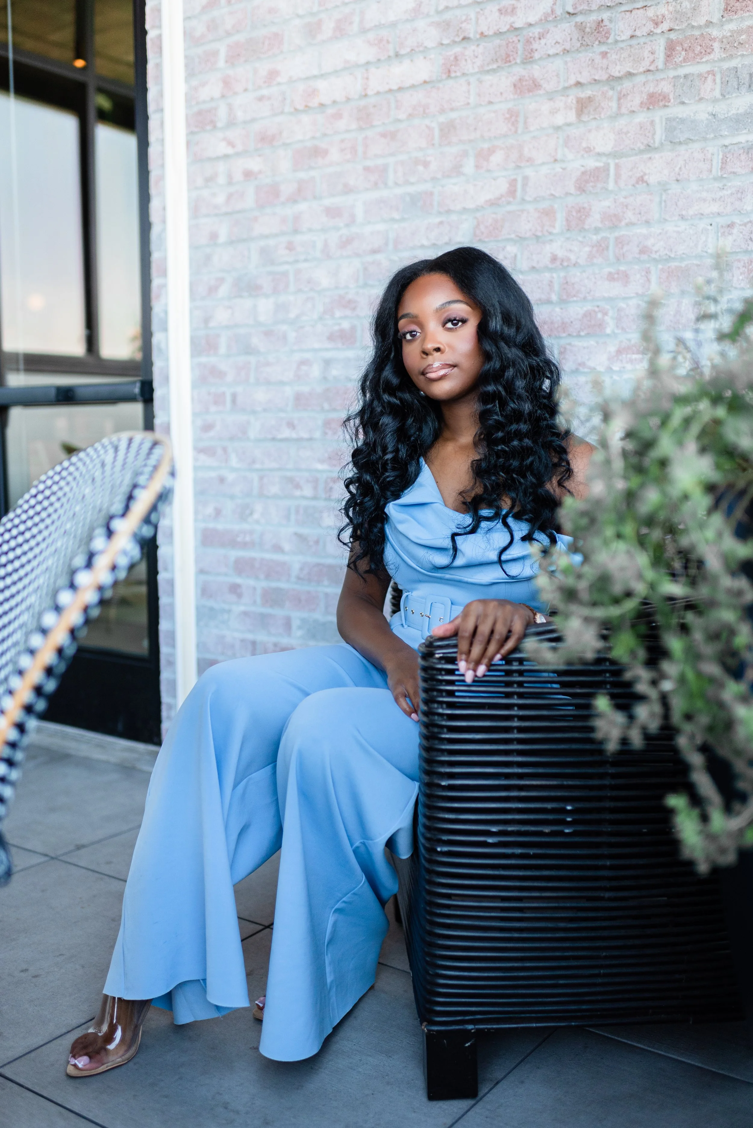A young woman with long, curly black hair wears a light blue dress and sits on a black chair, outdoors near a brick wall and a potted plant.
