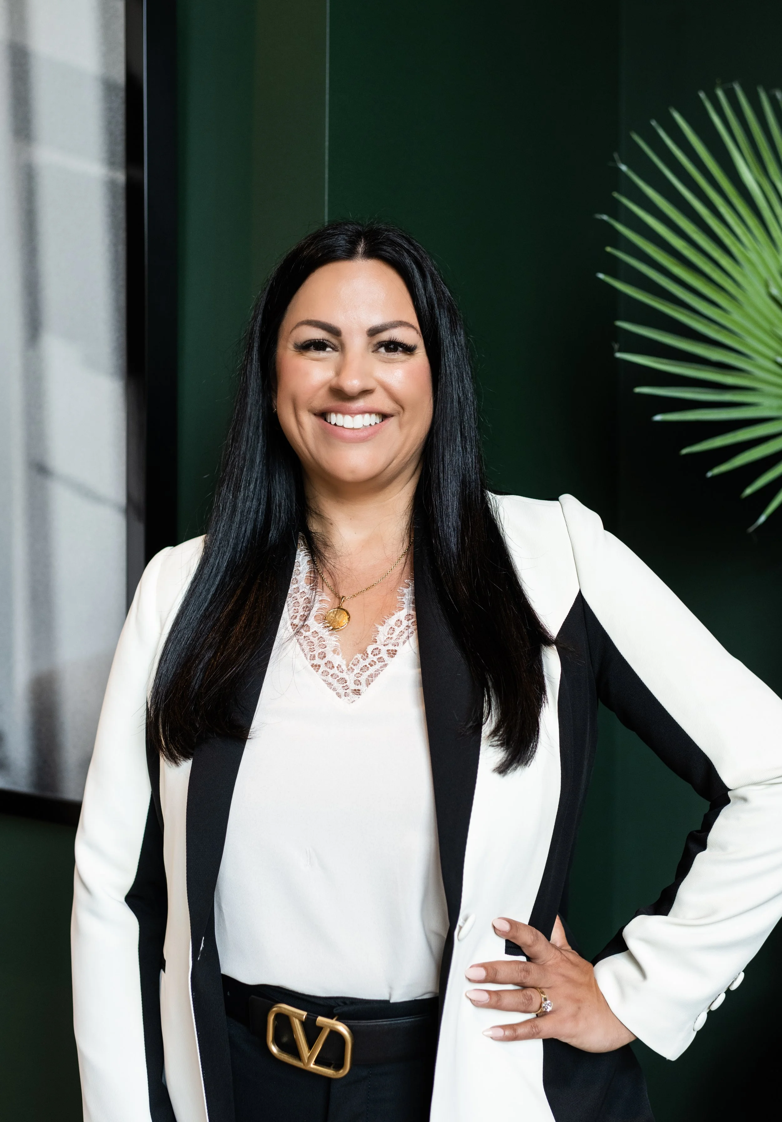 A woman with long black hair wearing a white blazer with black accents and a white lace top, standing in front of a dark green wall and large plant, smiling with one hand on her hip.
