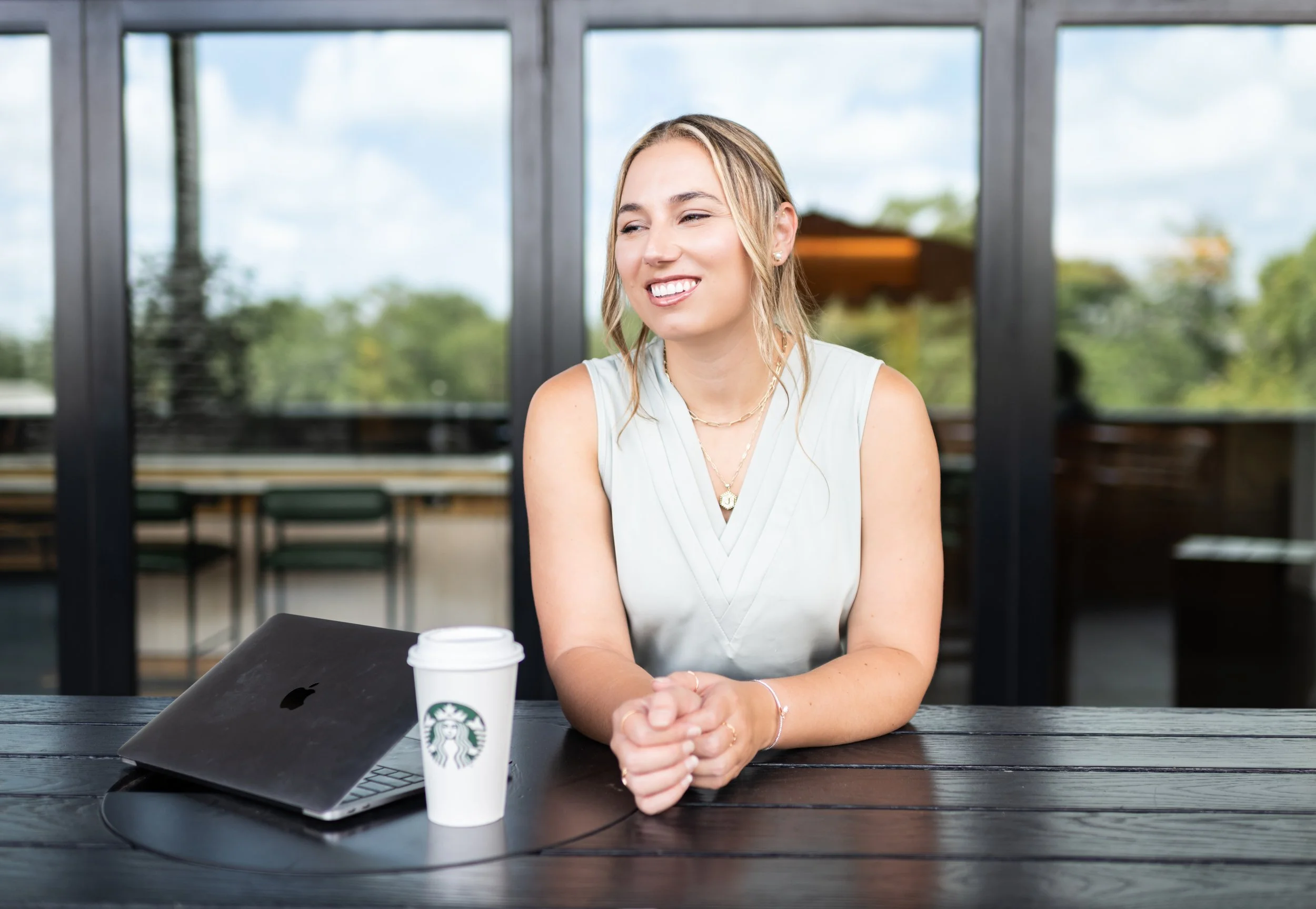 A smiling woman sitting at a dark wooden table with a closed MacBook and a Starbucks coffee cup, in a room with large windows showing a green outdoor scene.
