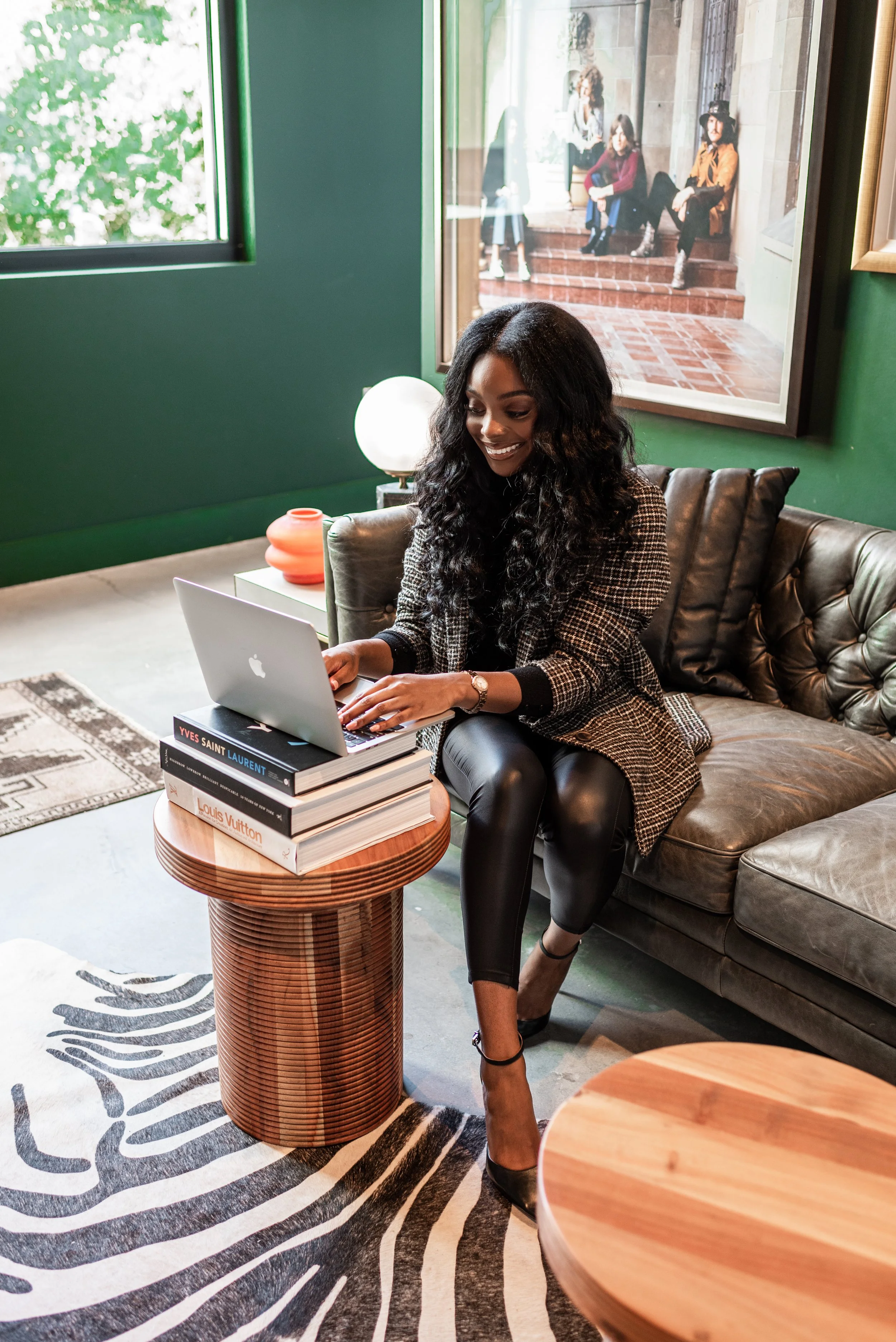 A woman with long, curly black hair working on a laptop in a cozy, modern living room with green walls, sitting on a leather sofa. She has a stack of fashion books on a round wooden table next to her, and a black and white zebra striped rug on the fl