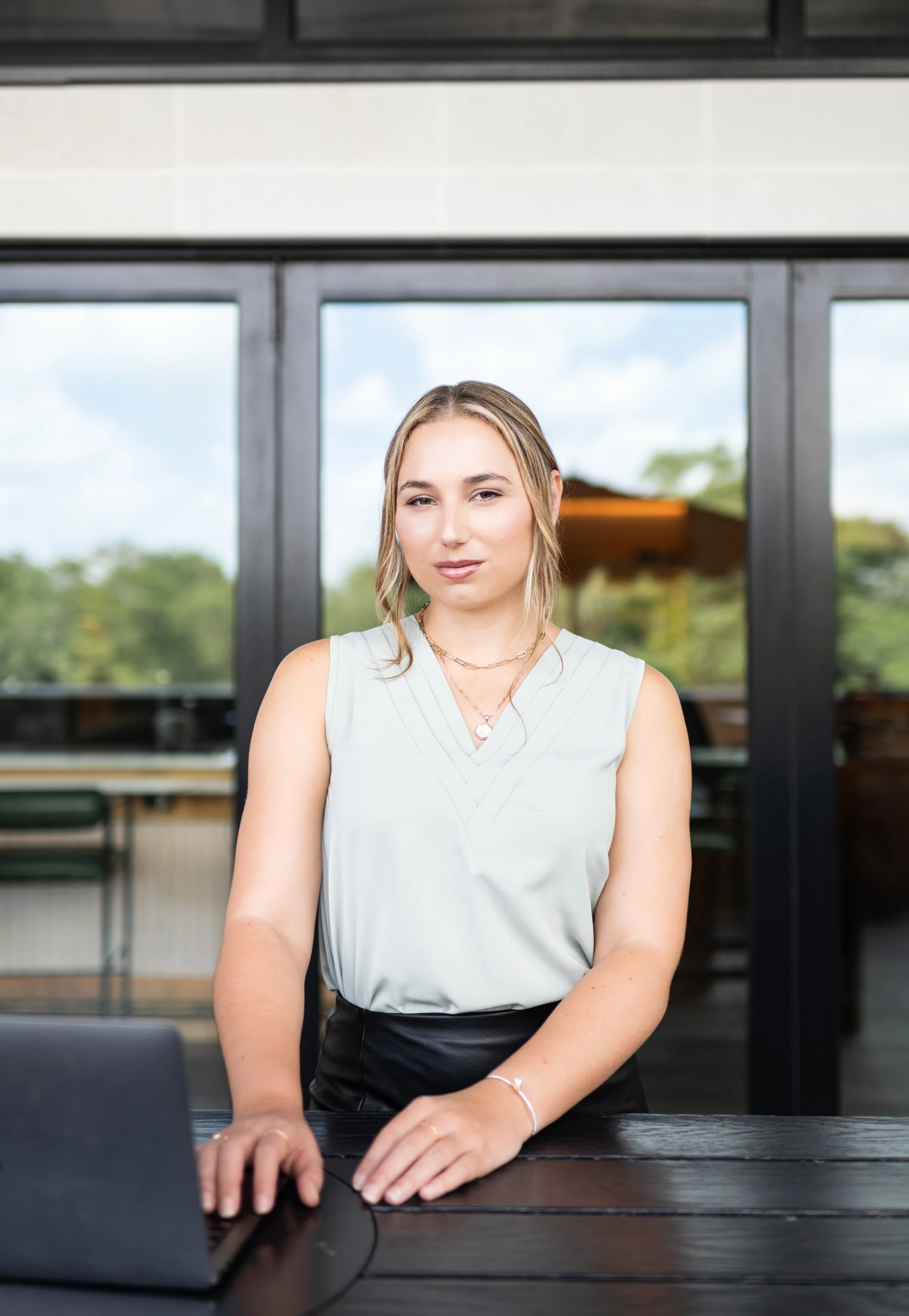 A young woman with blonde hair sitting at a desk with a laptop, inside a building with large windows showing a cloudy sky and green trees outside.
