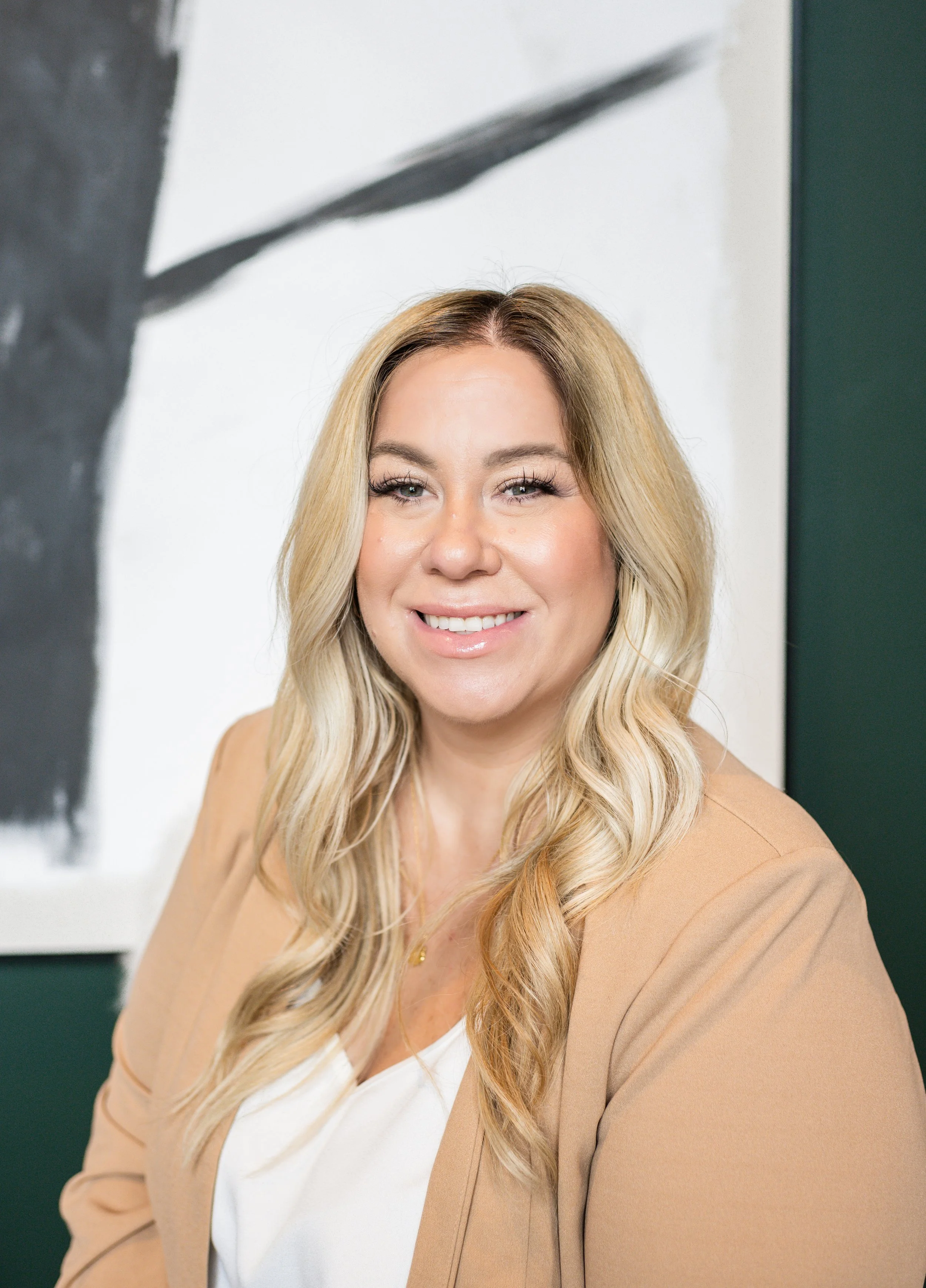 Portrait of a smiling woman with long blonde hair, wearing a beige blazer over a white top, standing indoors in front of a white wall with black and green abstract art.