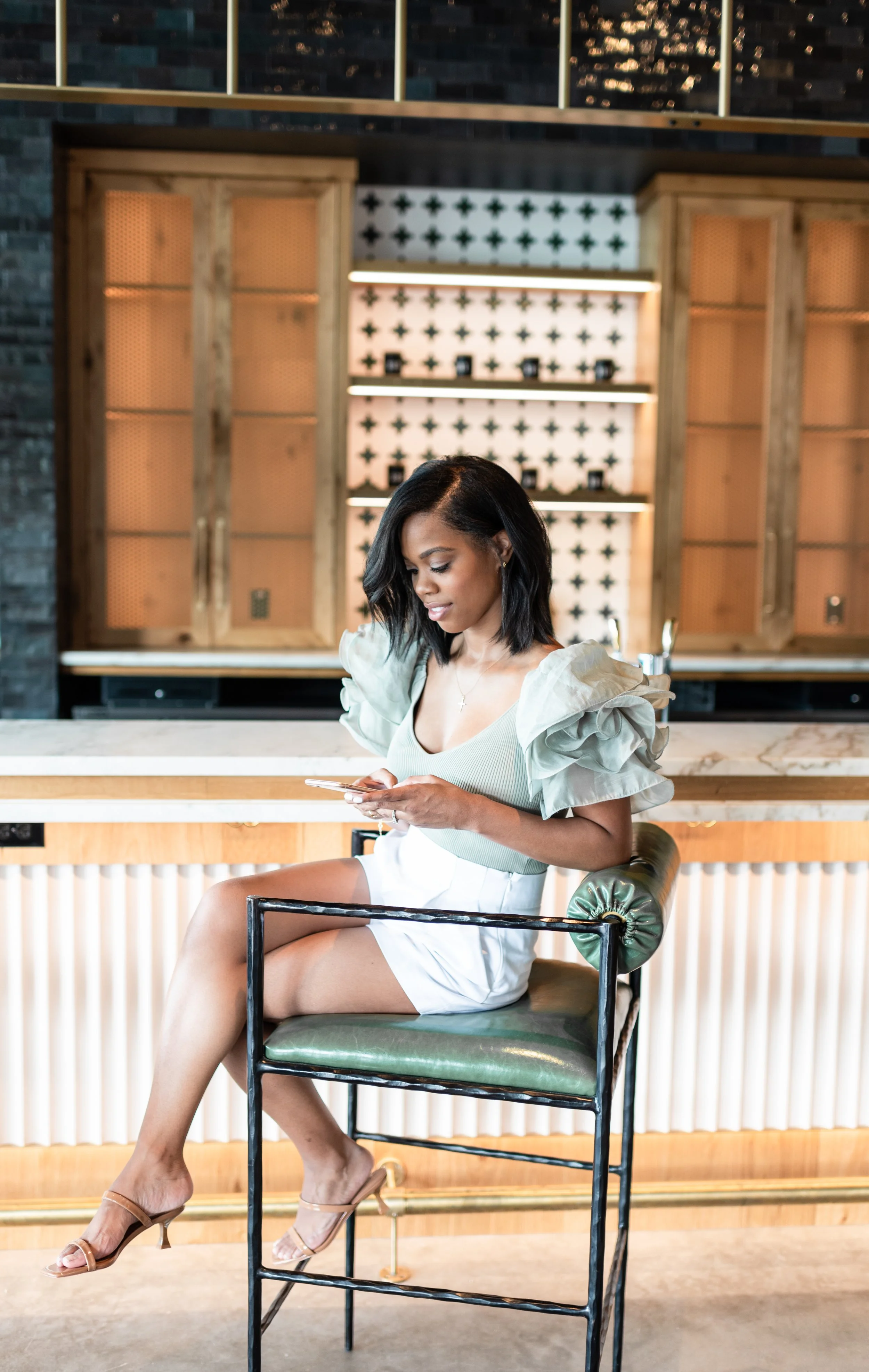 A woman with shoulder-length black hair, wearing a light green top with ruffled sleeves and white shorts, sitting on a green cushioned chair at a bar, looking at her phone.