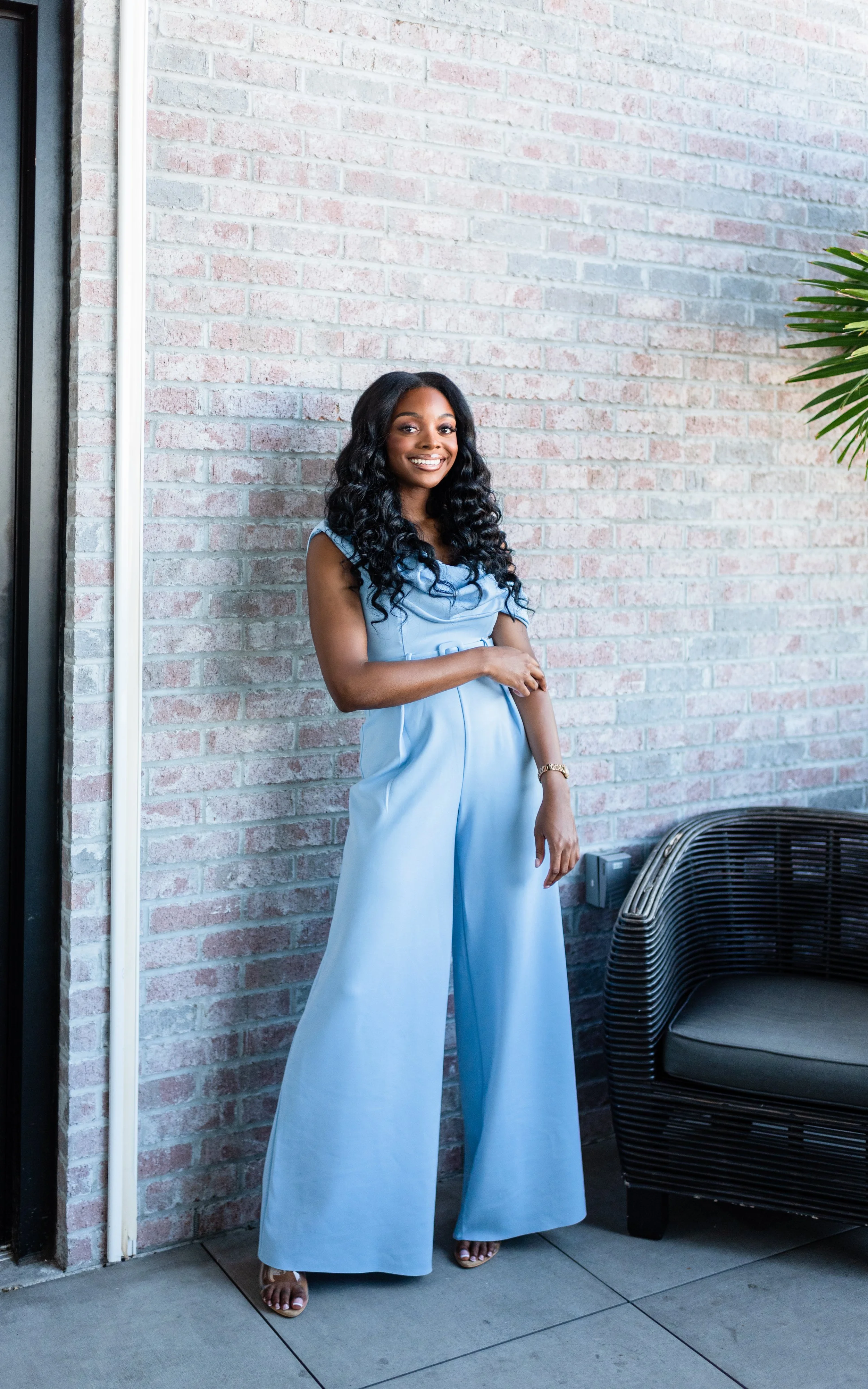 A woman in a light blue jumpsuit with wide-leg pants and a smiling face stands beside a brick wall. She has black curly hair and wears a wristwatch. There is wicker outdoor furniture and a potted plant nearby.