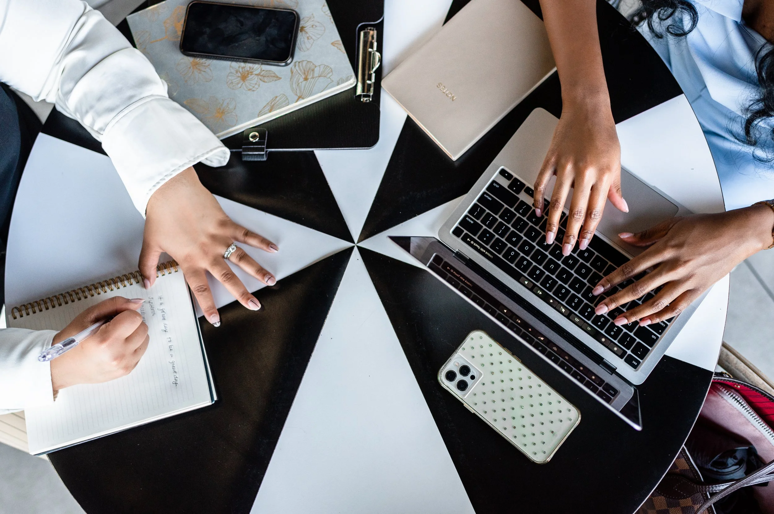Top-down view of two people working at a round black and white table with laptops, notebooks, a smartphone, and a pen.