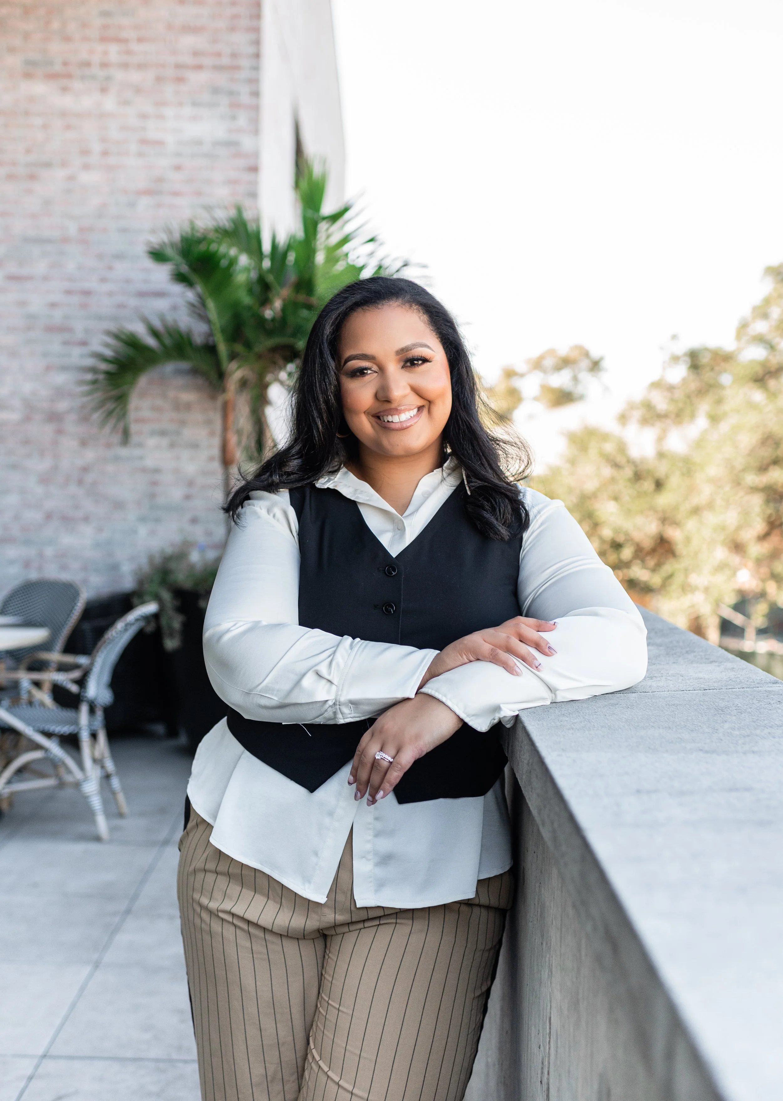 A woman with dark hair, smiling, leaning on a concrete railing, outdoors with trees and a brick wall in the background.