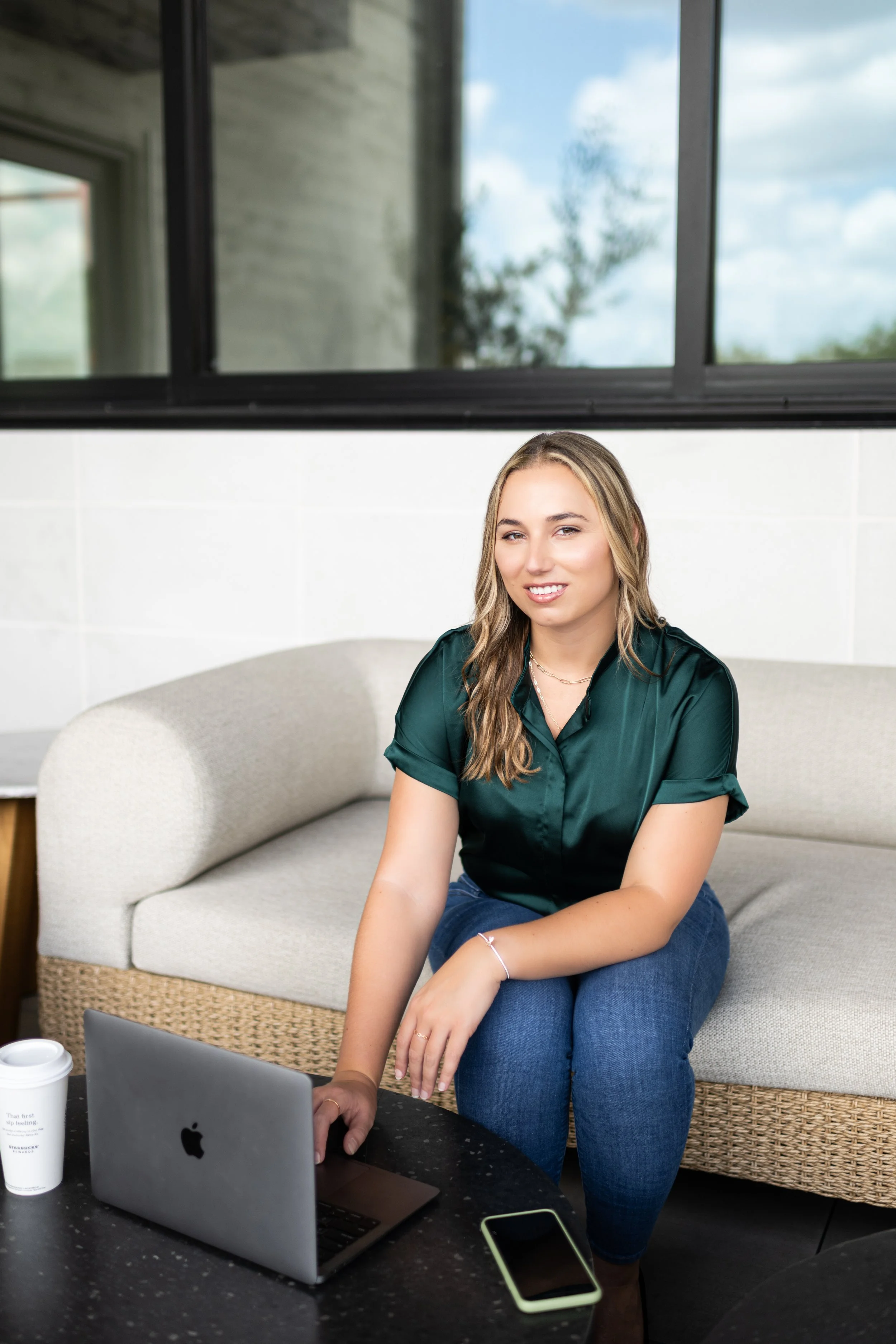 A woman sitting on a sofa with a laptop, smartphone, and coffee cup on a table in front of her, in a room with large windows showing a cloudy sky and trees outside.