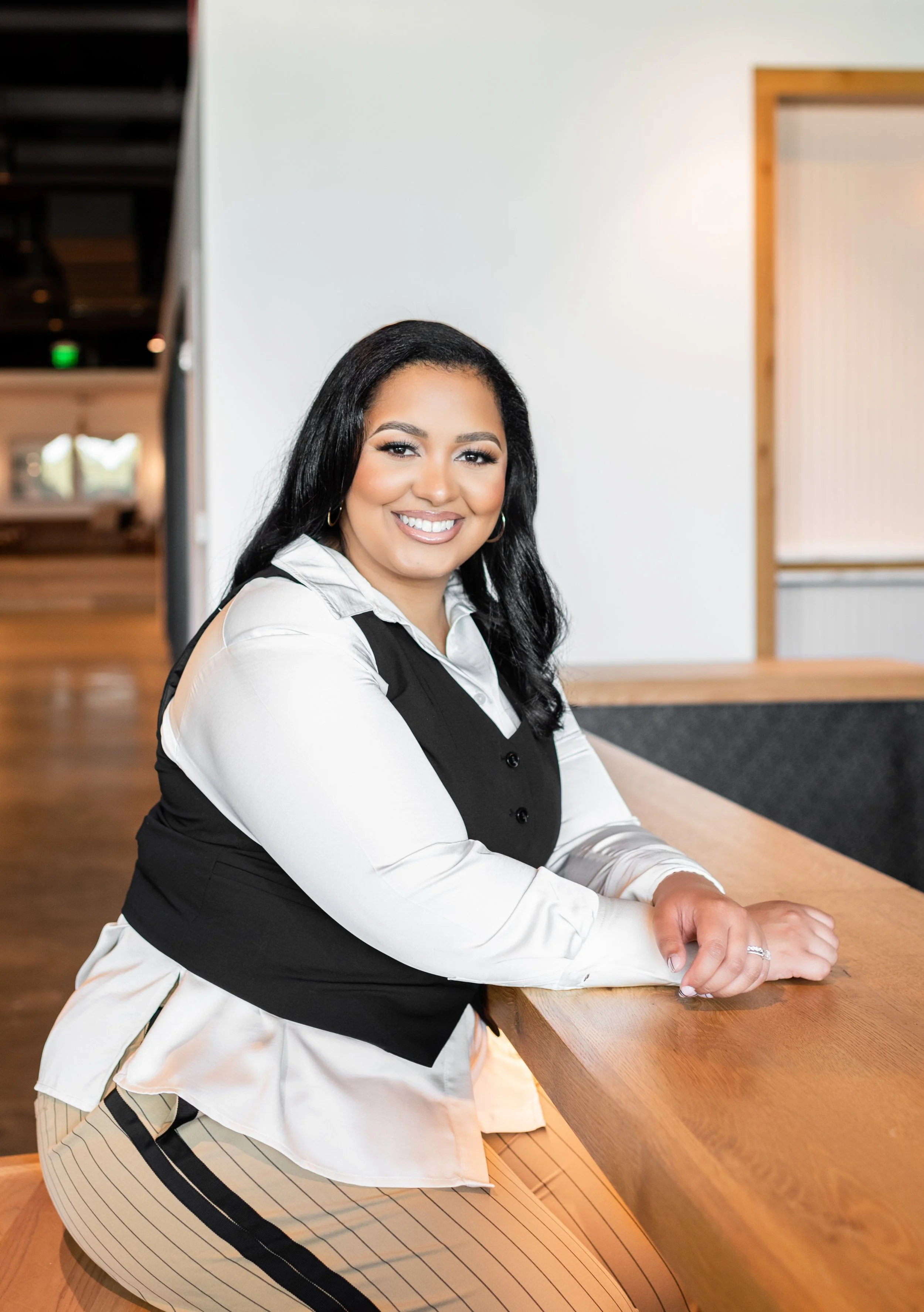 A woman with long black hair smiling, sitting at a wooden table in a modern indoor space.