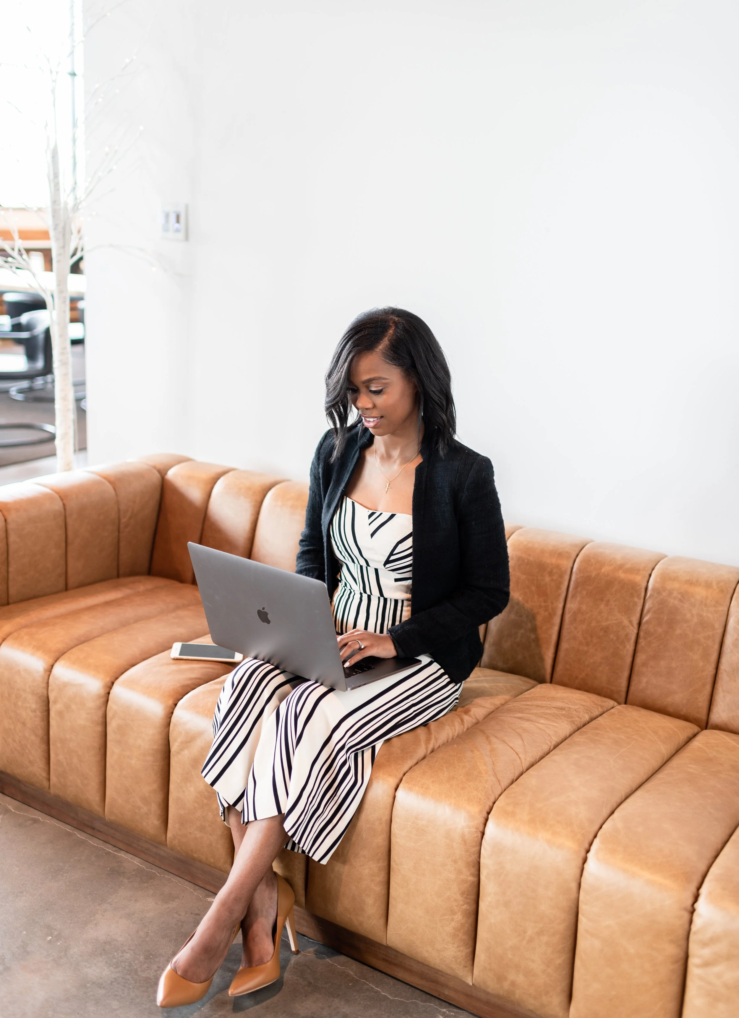 A woman sitting on a tan leather couch, working on a silver laptop with a smartphone nearby, in a bright, modern room.