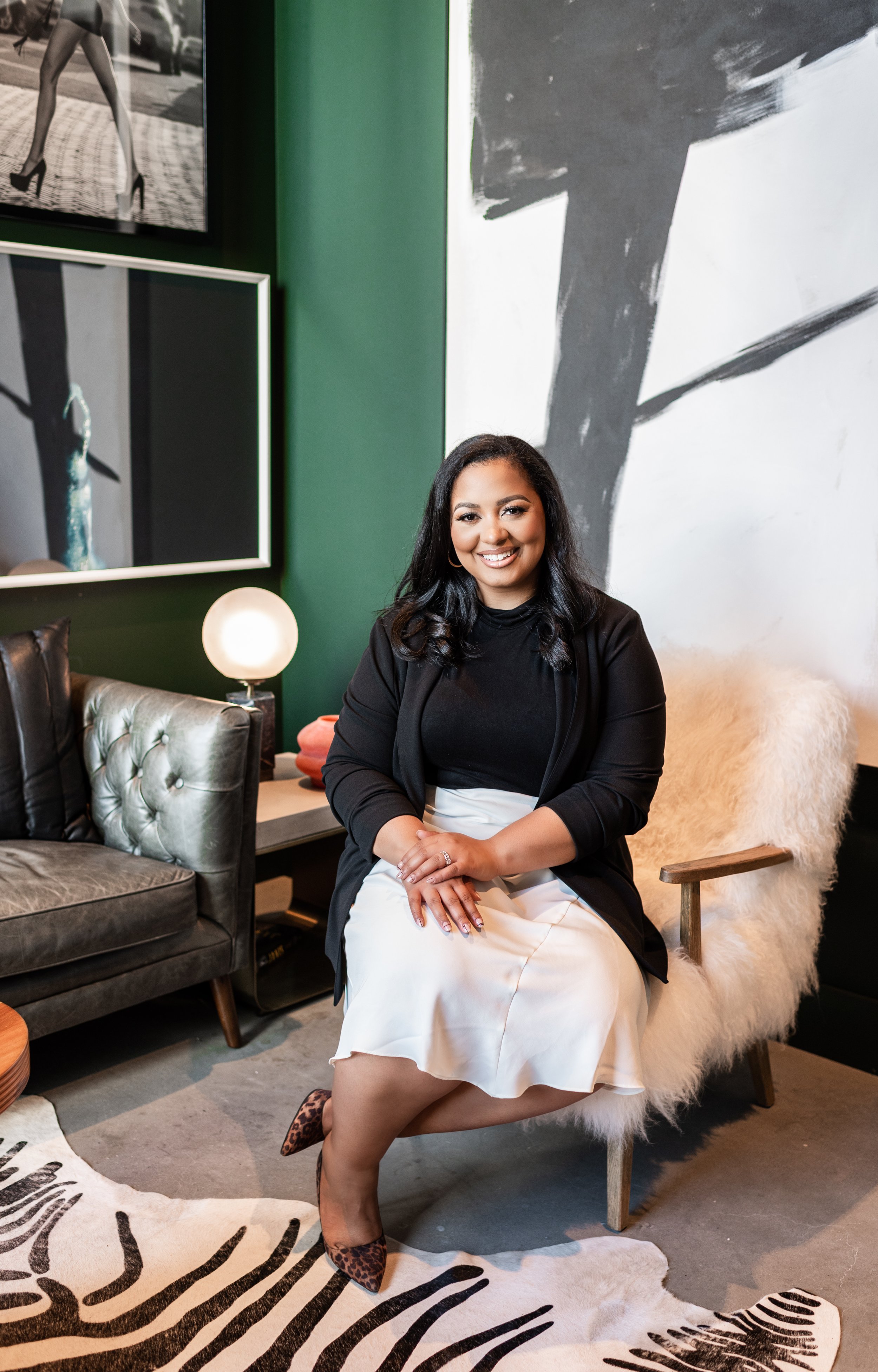 A woman with black hair sitting on a cozy, white, furry chair in a modern, stylish living room with black, white, and green decor, artwork, and a lamp.