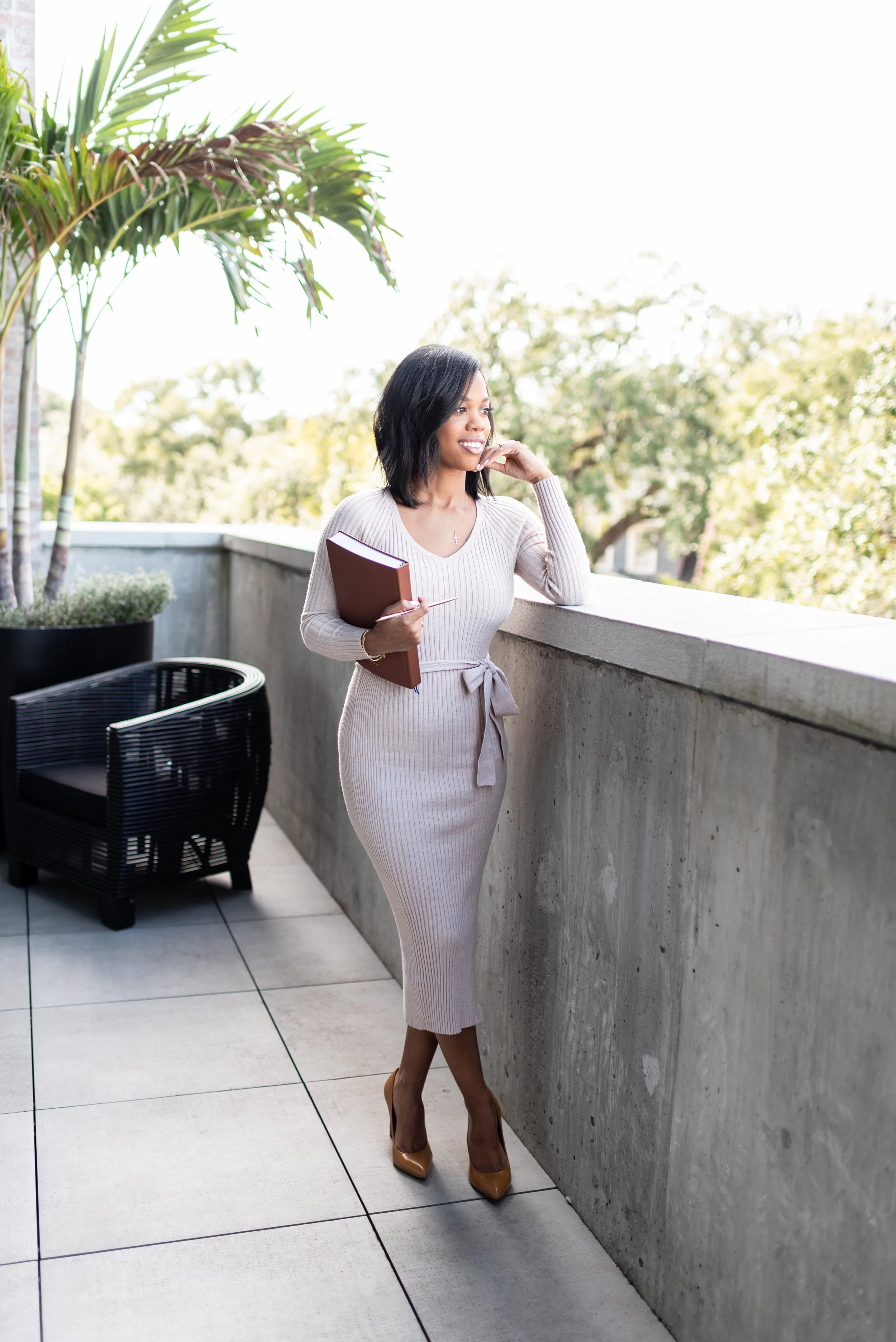 A woman in a beige ribbed dress, holding a brown notebook, stands on an outdoor balcony with a concrete railing, potted plants, and black wicker chairs, looking thoughtfully into the distance with trees in the background.
