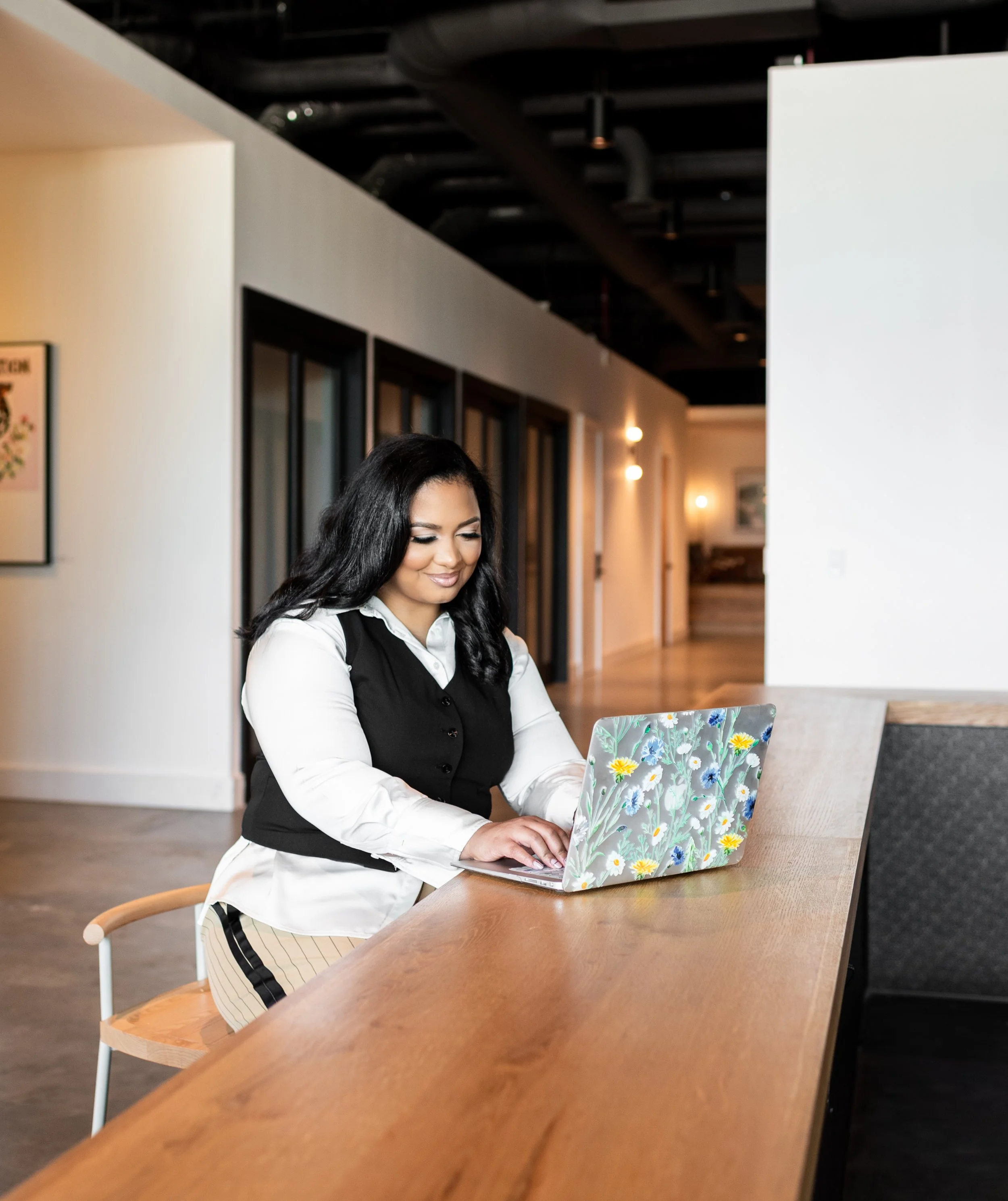 A woman with dark, shoulder-length hair working on a laptop with a floral design cover in a modern, well-lit space.