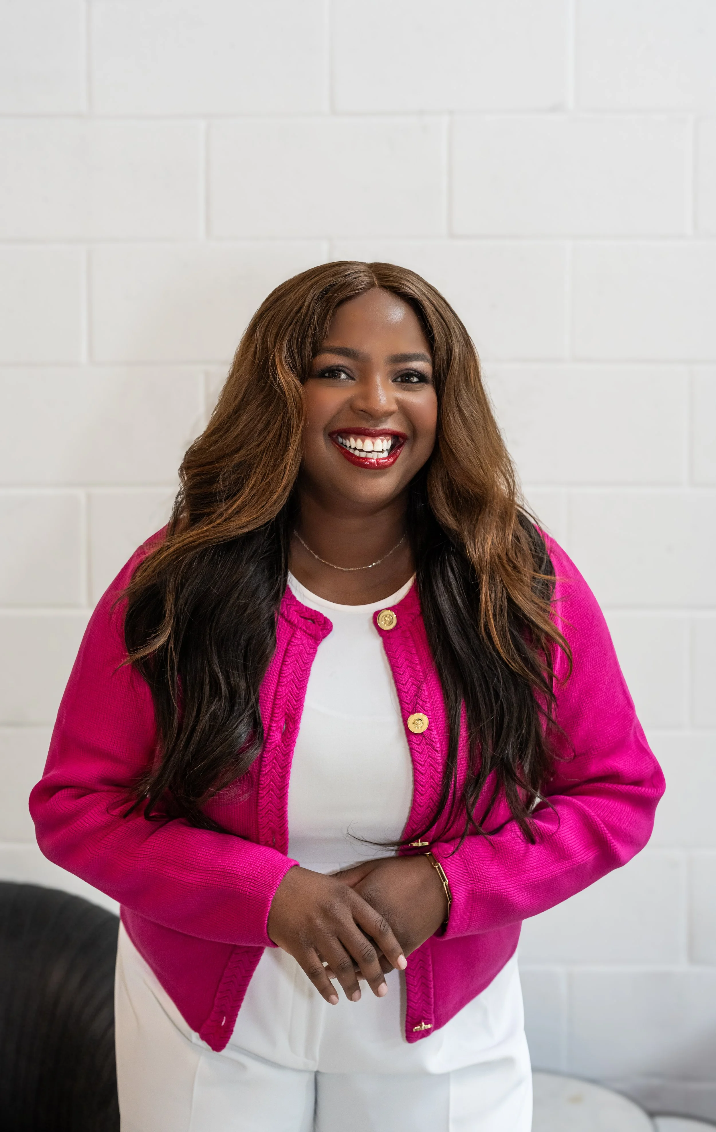 A woman with long wavy hair, wearing a pink jacket and white top, smiling and standing against a white brick wall.