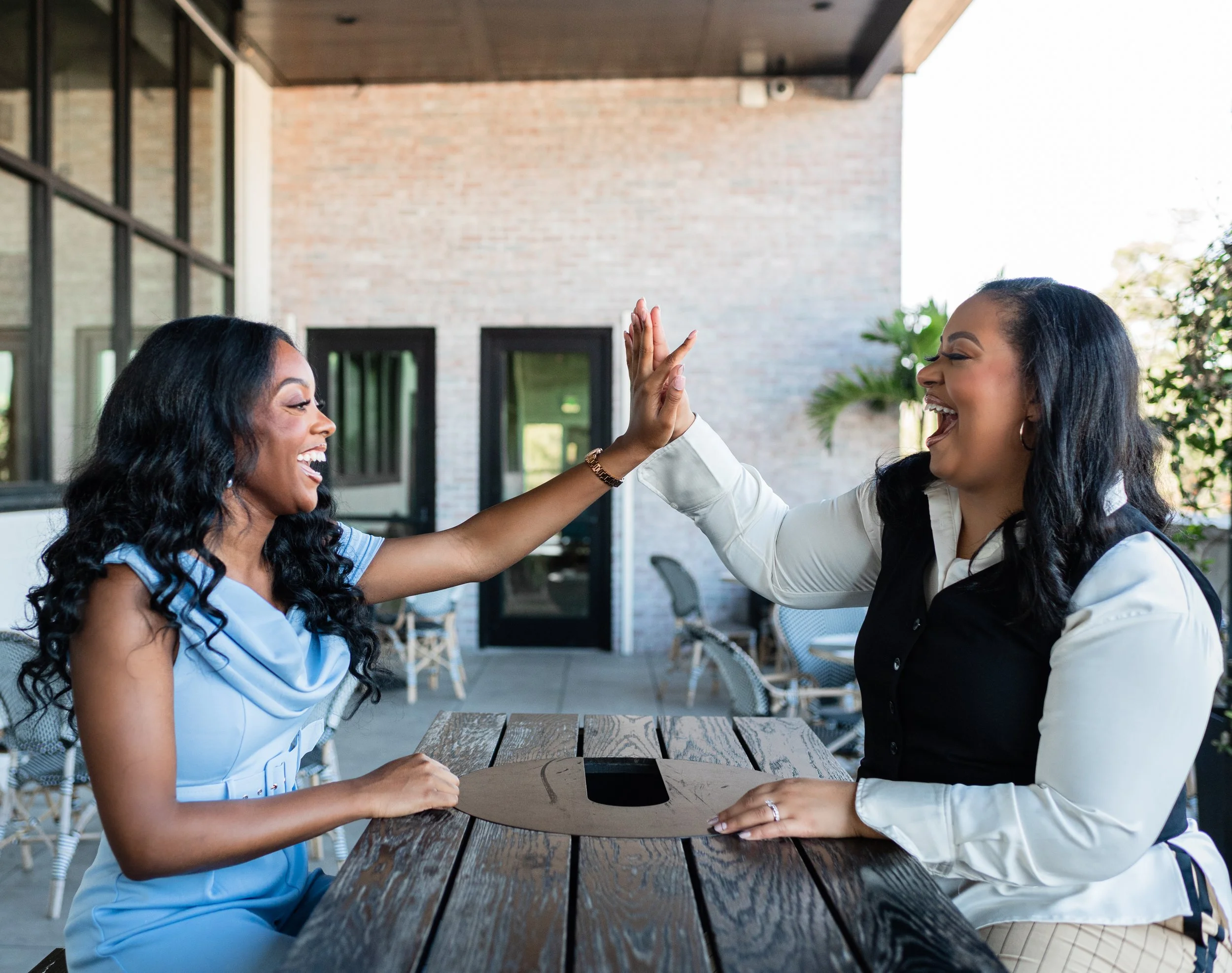 Two women sitting at a table outdoors giving each other a high-five, smiling and laughing, with a brick building and windows in the background.
