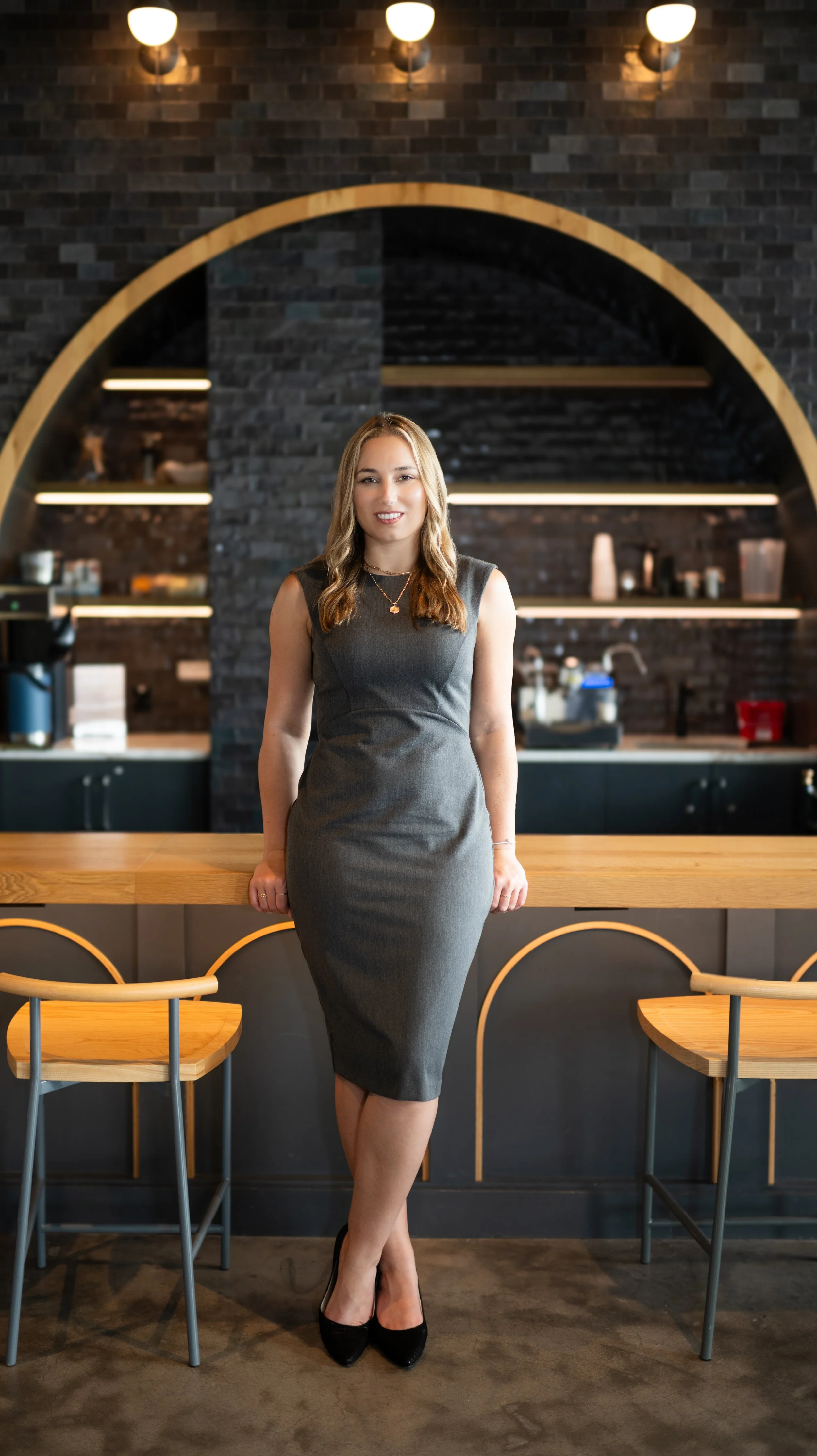 A woman in a gray sleeveless dress and black heels standing in a modern cafe with bar stools, a wooden counter, and a black brick wall with shelves in the background.