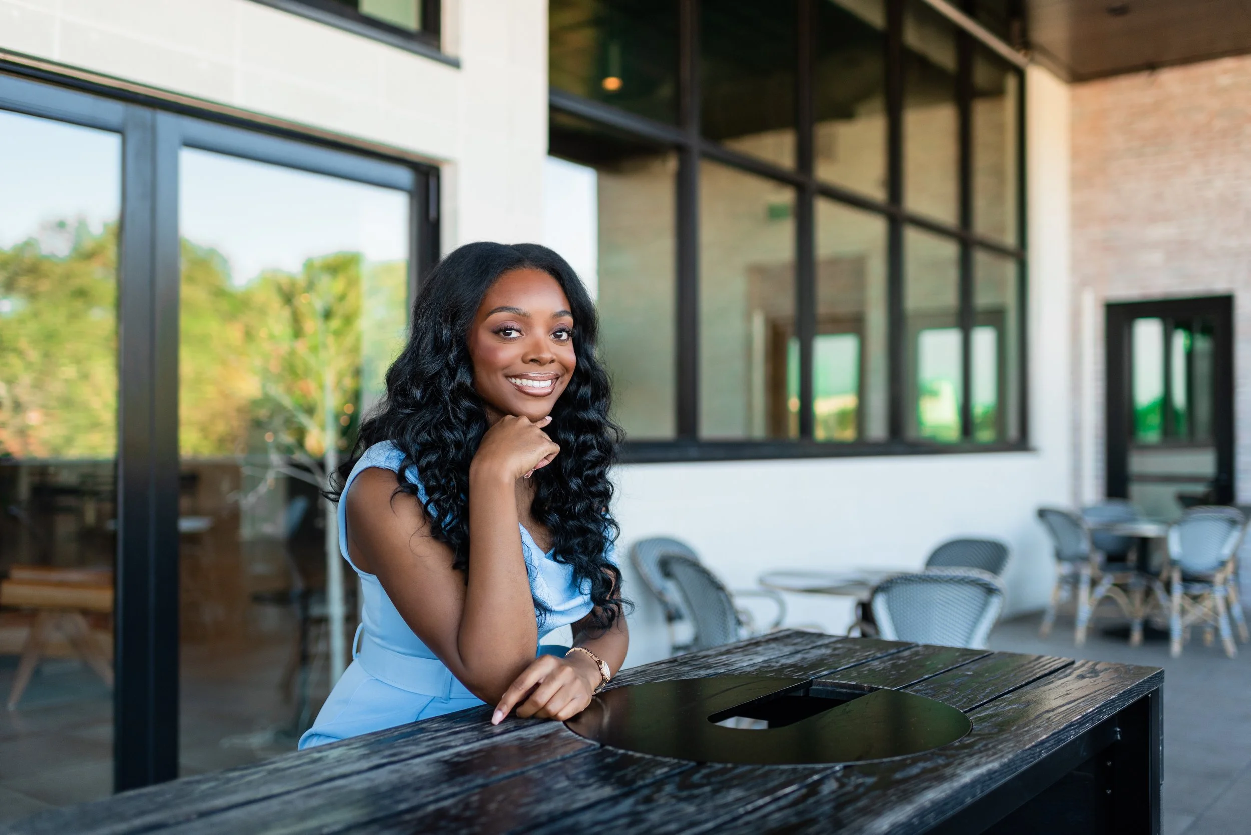 A smiling woman with long, curly black hair sitting at a dark wooden table outside a modern building with large glass windows.