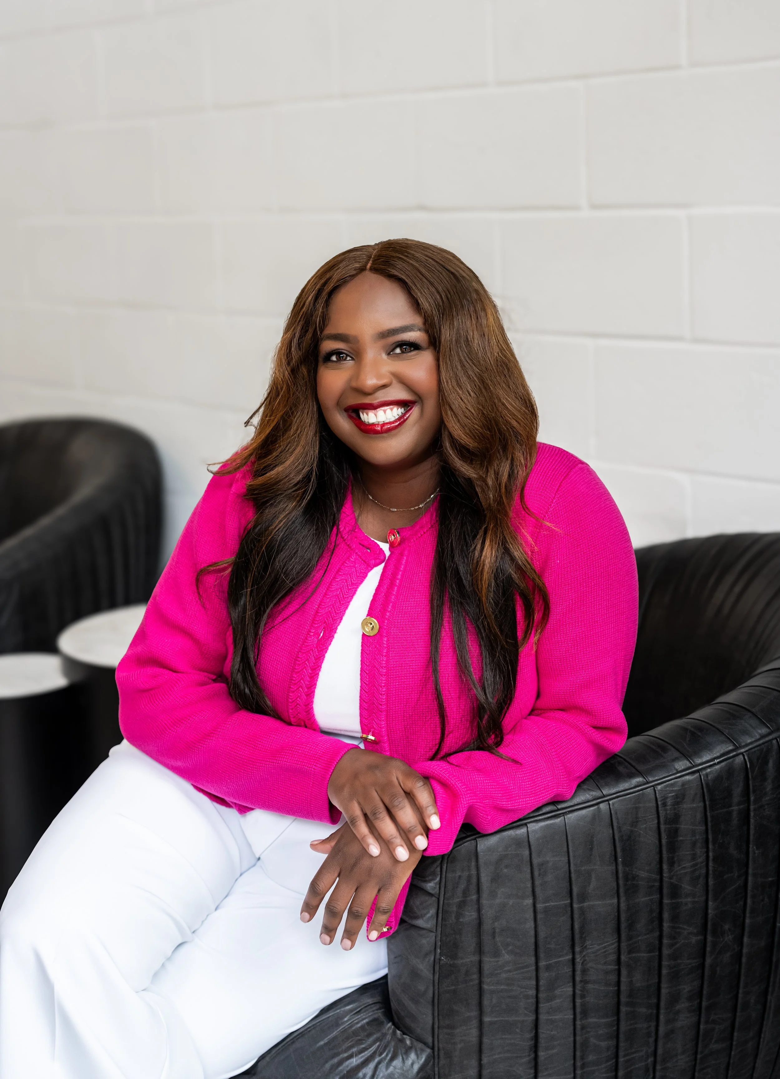 A woman with dark brown skin and long wavy hair, wearing a bright pink jacket and white pants, sitting on a black leather couch with a white brick wall behind her, smiling at the camera.