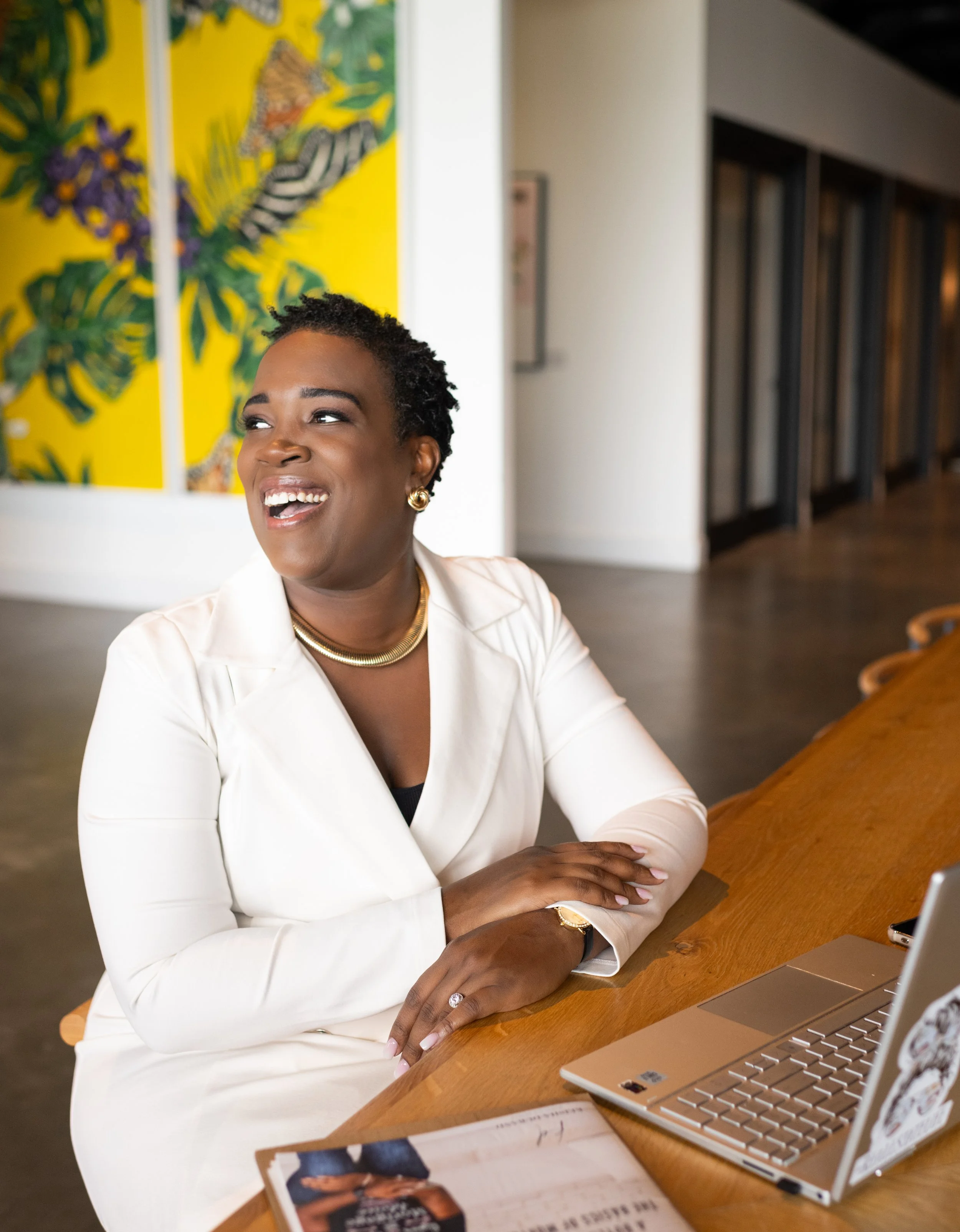 A woman sitting at a wooden table with a laptop, smiling and looking to her right, in a modern indoor space with colorful artwork on the wall behind her.