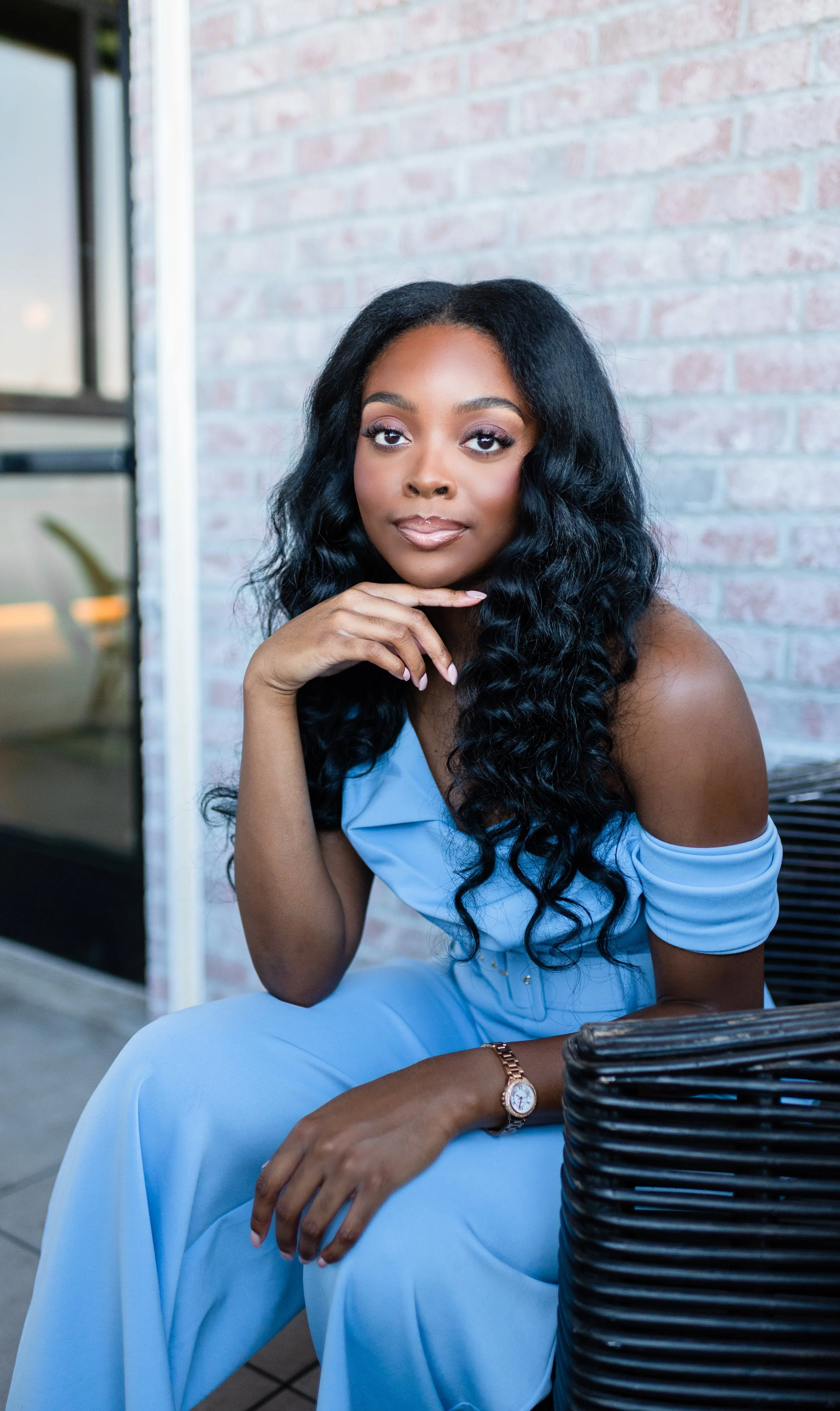 A woman with long black curly hair, wearing a light blue off-shoulder dress, sitting on a wicker chair outside by a brick wall, posing with her hand under her chin, wearing a watch on her left wrist.