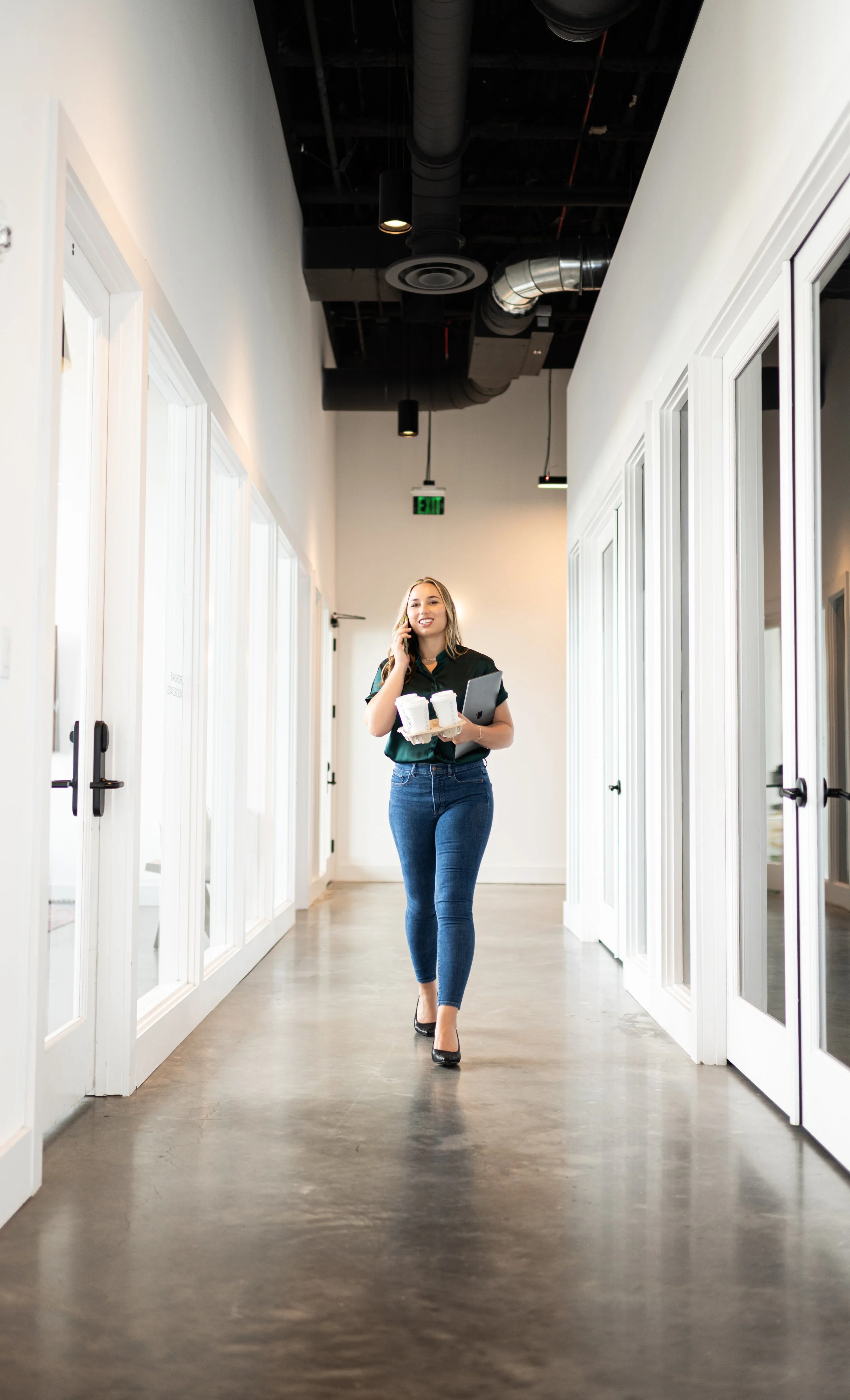 A woman walking down an office hallway talking on her cell phone, carrying two coffee cups and a laptop.