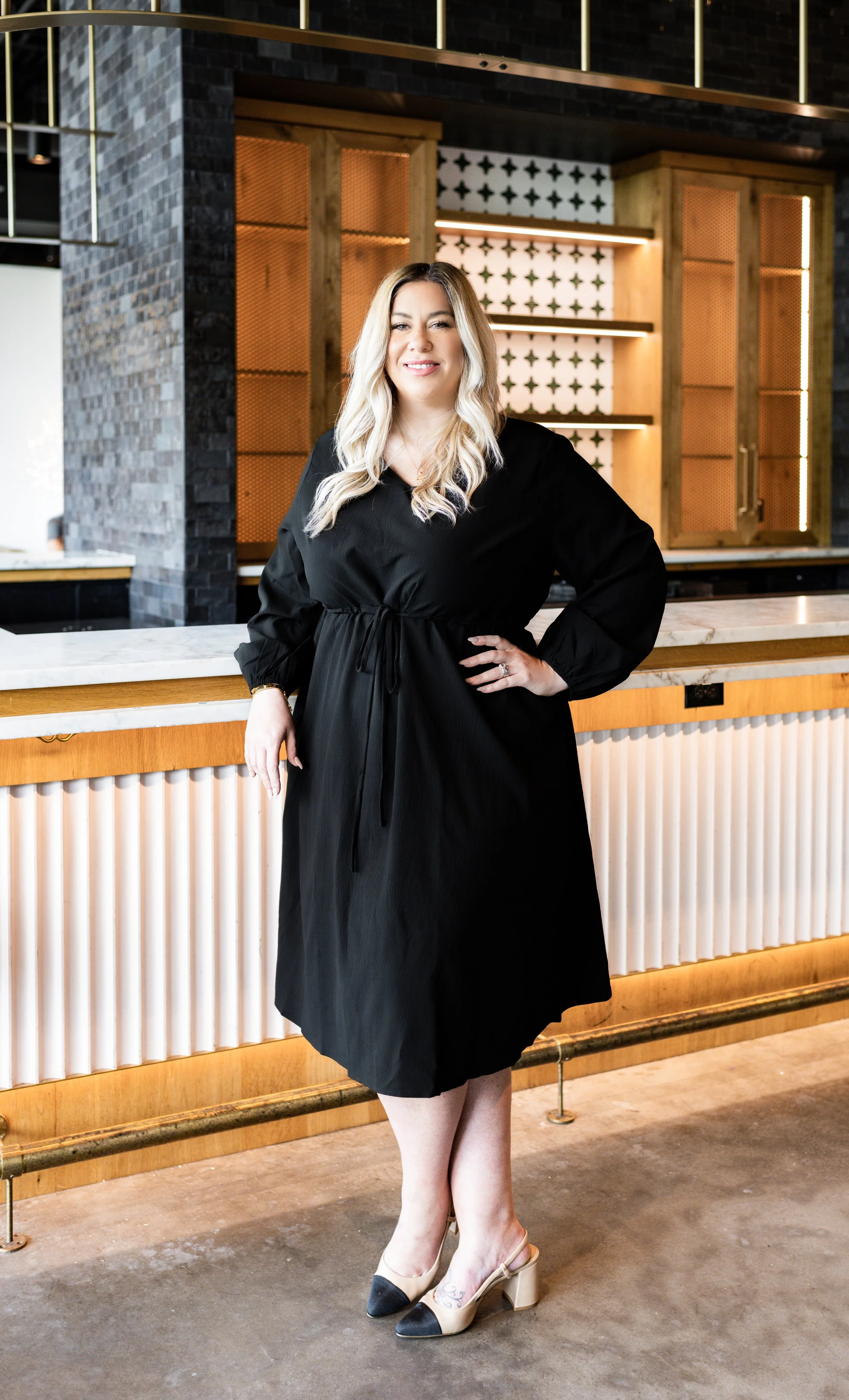 A woman with long blonde hair standing at a bar, wearing a black dress and two-tone high heels, smiling at the camera in a modern decor setting.