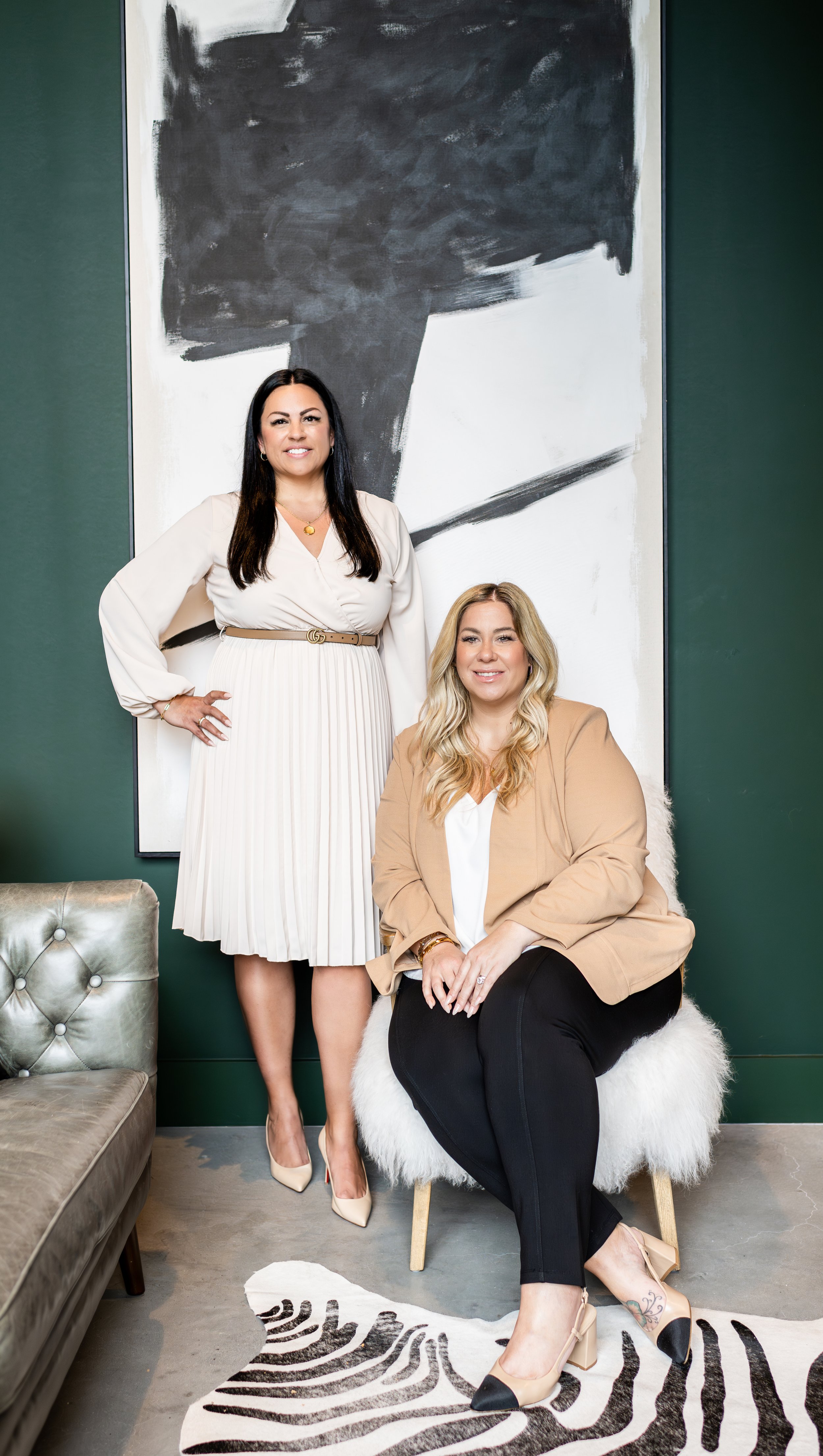 Two women in professional attire pose for a photo in an interior setting with modern decor and abstract wall art.