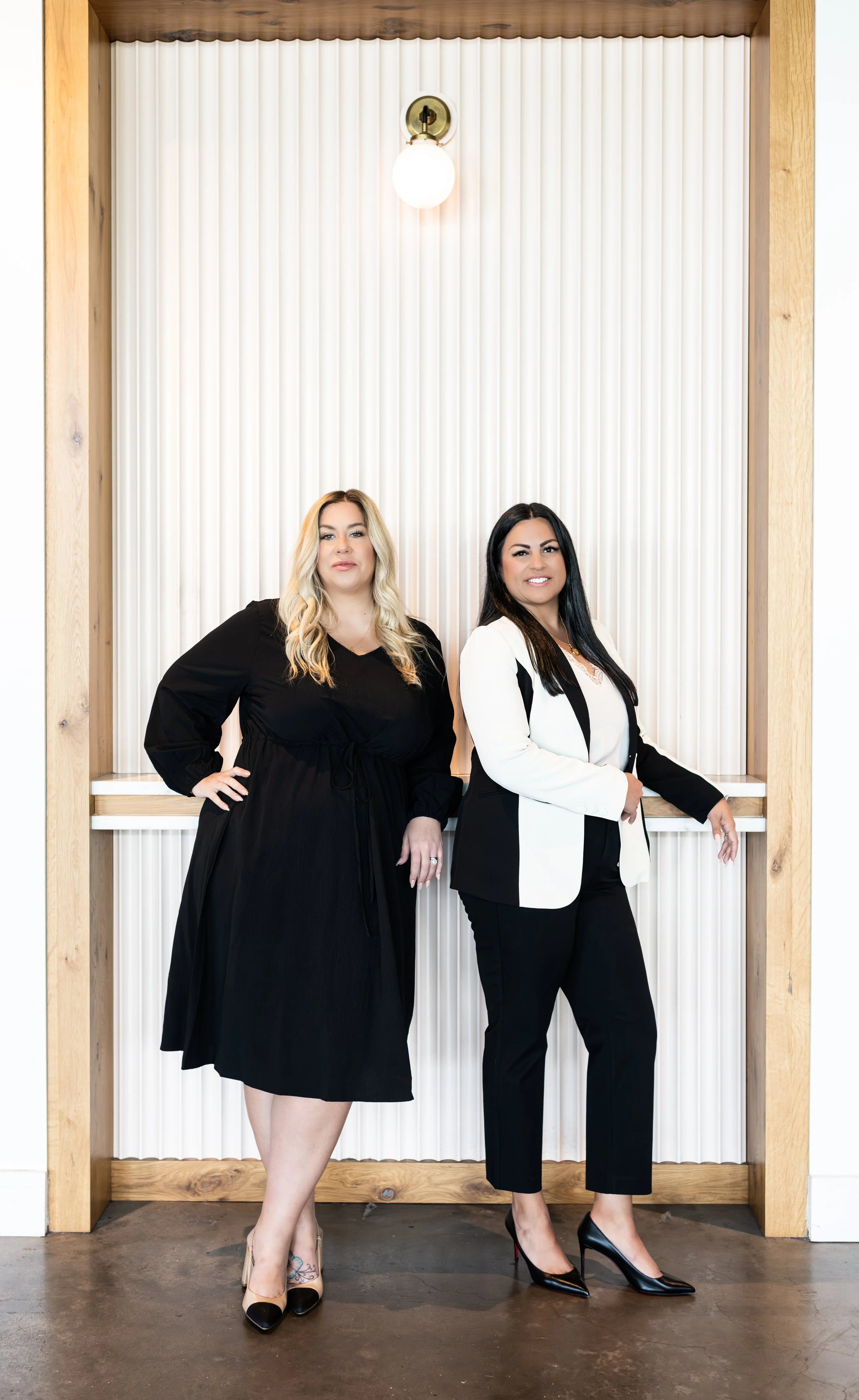 Two women standing in front of a textured white wall with wooden framing and a light fixture, dressed in business attire, looking at the camera.