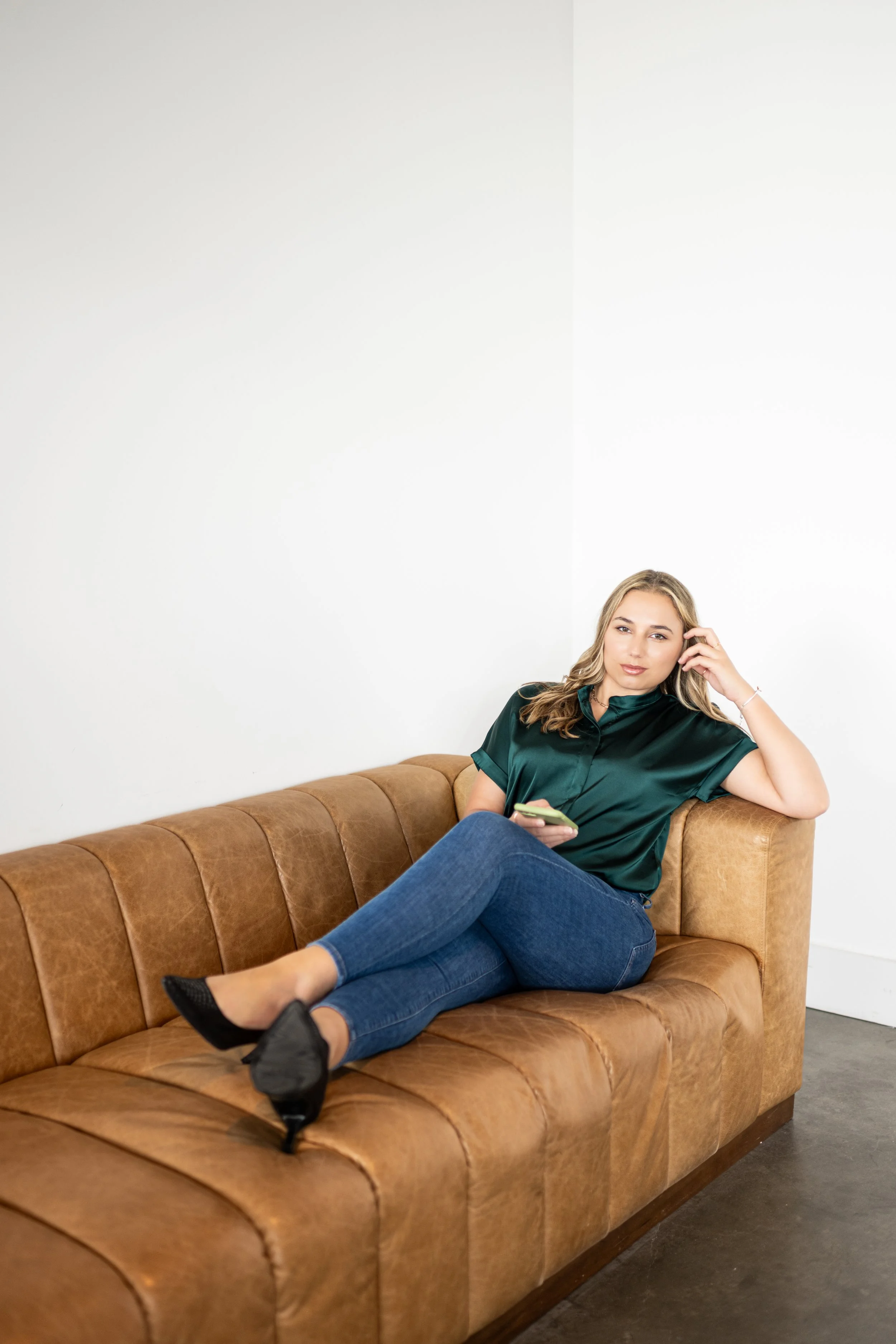 A young woman sitting on a brown leather couch, holding a smartphone, with her legs crossed, in a room with white walls.