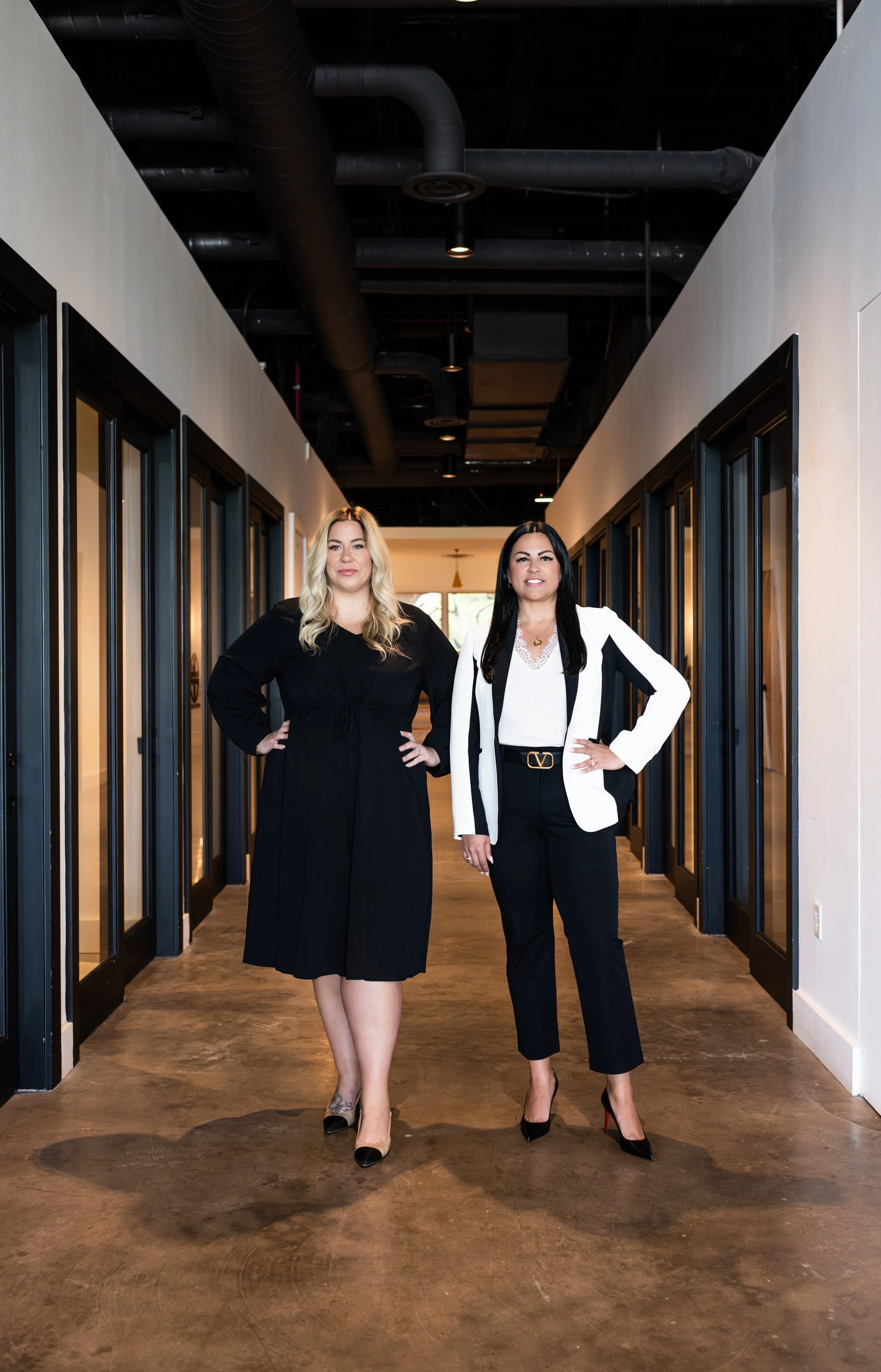 Two women standing confidently in a modern indoor corridor, one in a black dress and the other in a white blazer and black pants, both with hands on hips.