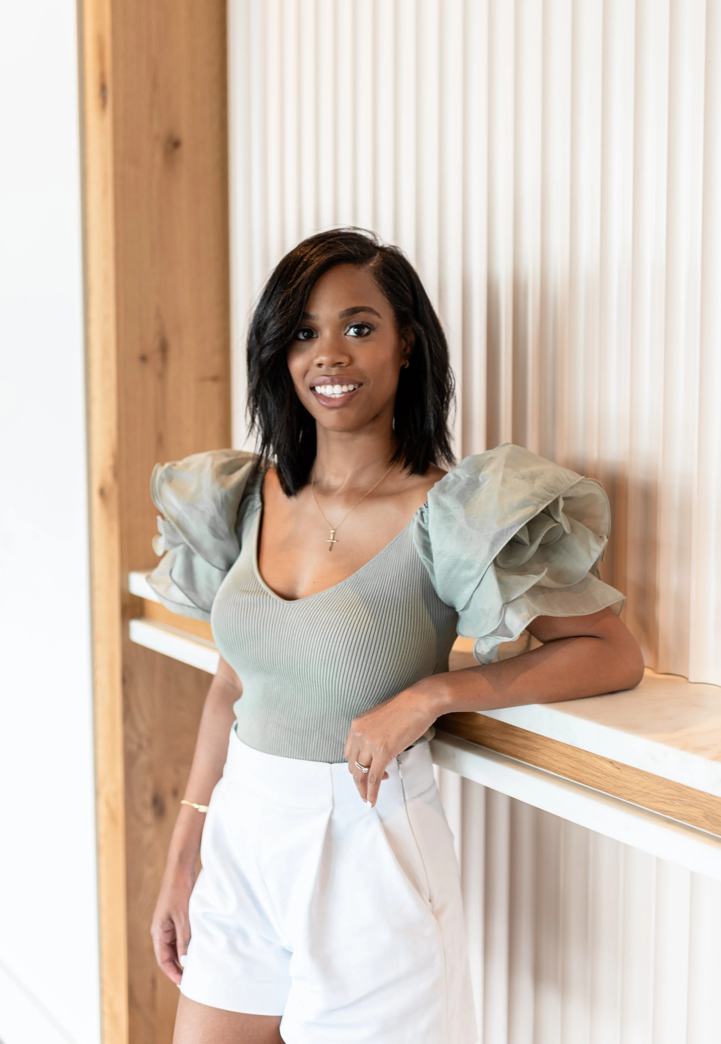 A young woman with dark hair and a smile, wearing a light green top with puffed sleeves and white shorts, leaning against a wooden and marble wall.