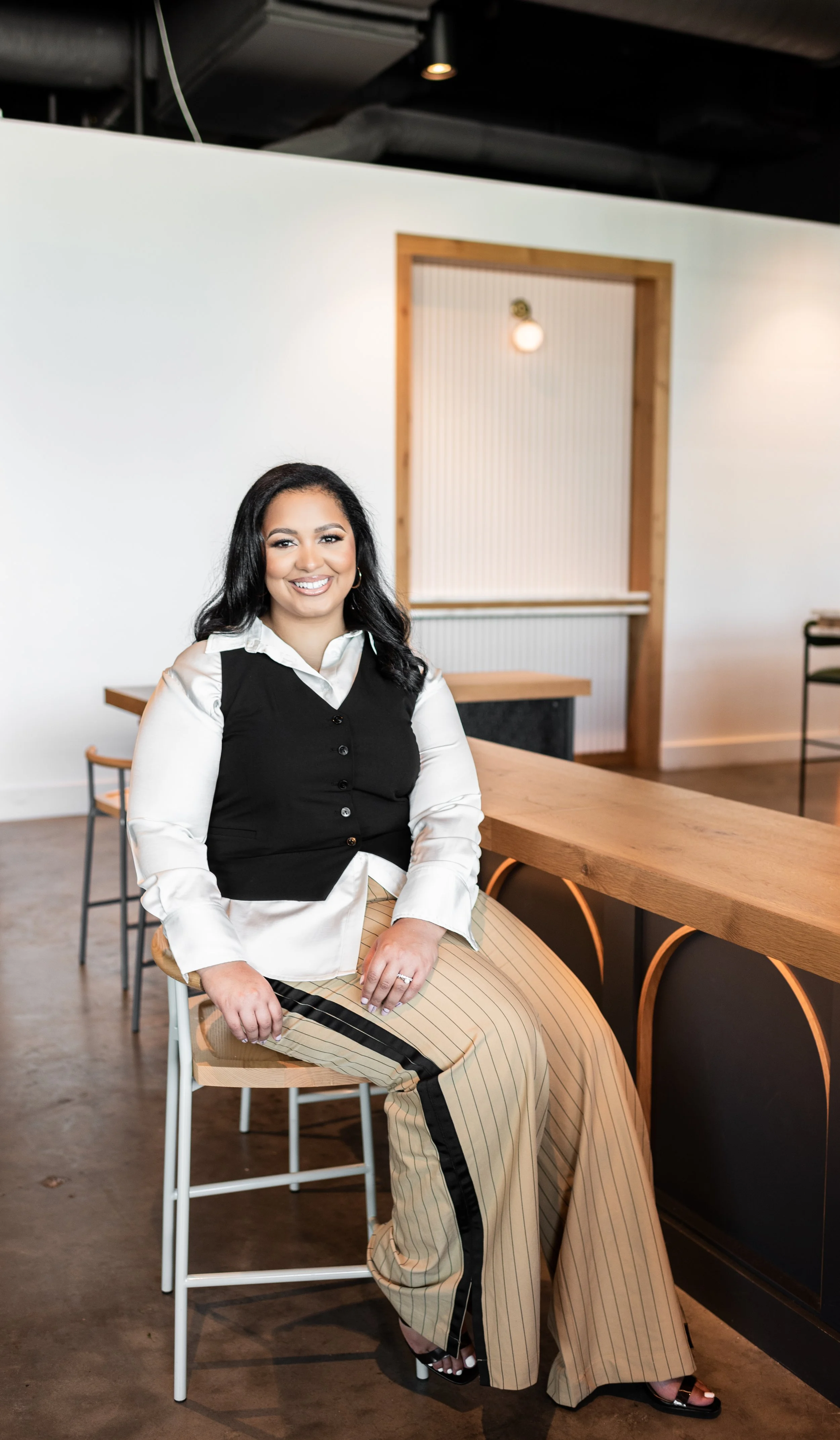 A woman sitting on a barstool in a modern interior space, smiling at the camera, wearing a white shirt, black vest, striped beige and black wide-leg pants, with dark hair styled in loose waves.