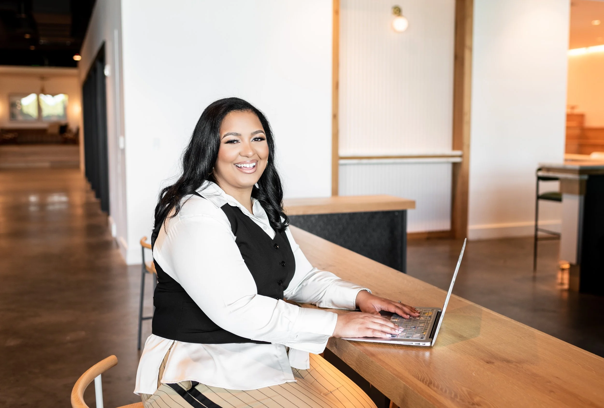 A woman with long black hair, smiling while using a laptop at a wooden table in a modern, well-lit interior space.