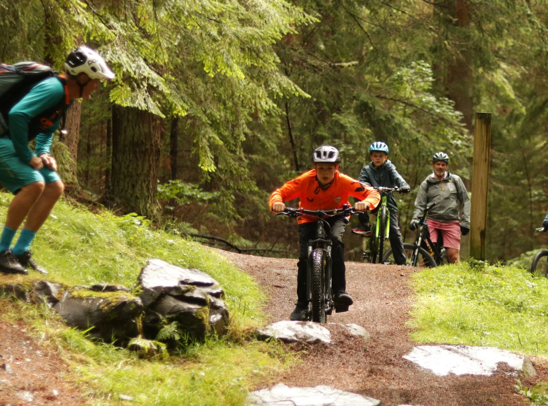 A group of children and adults mountain biking on a trail through a lush, green forest. One child in an orange jacket is riding a bike on the dirt path, while three others wait behind. An adult is to the right, and another adult with a helmet is on the far left, observing the scene.