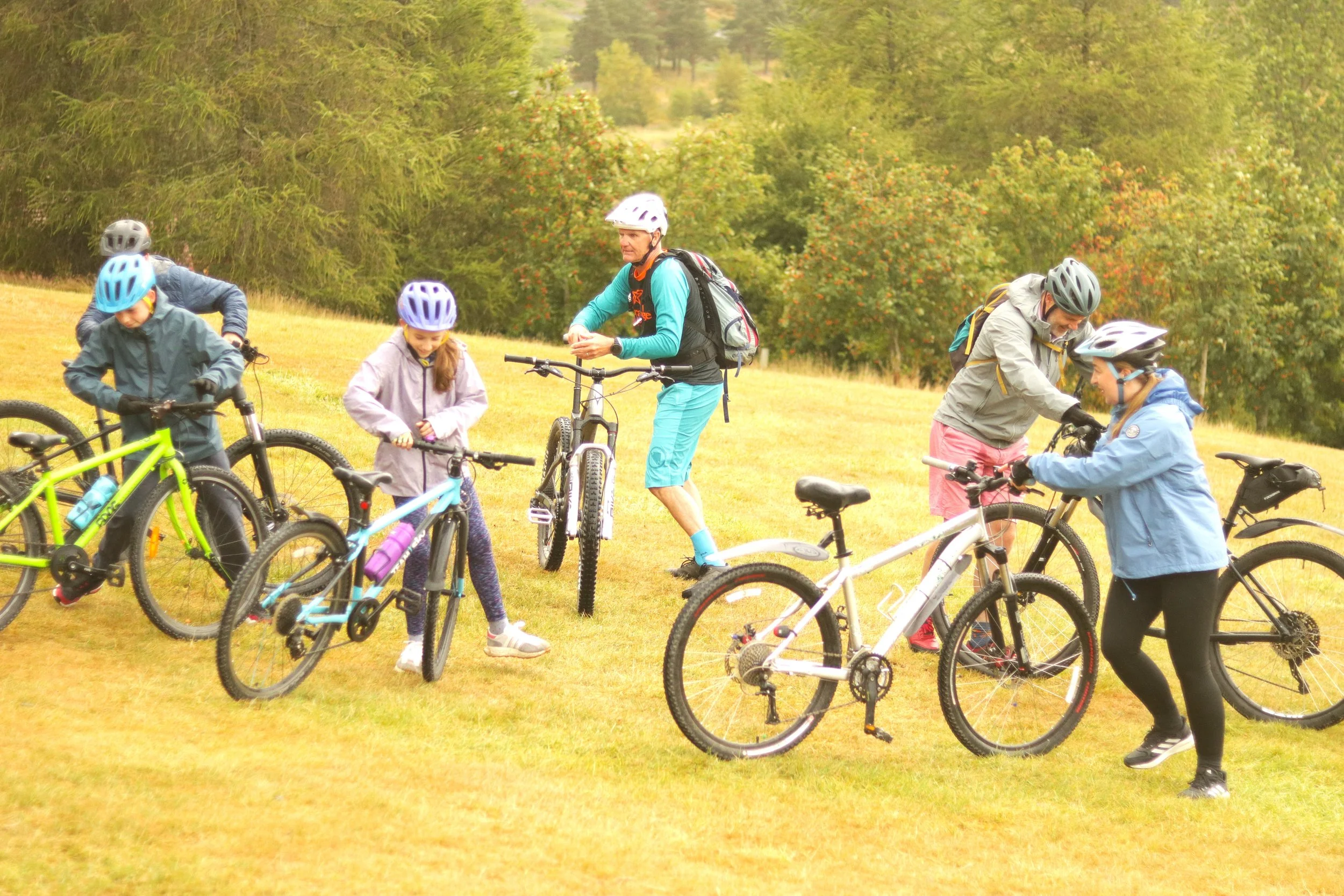 A group of children and adults with helmets and bicycles gathered on a grassy area outdoors, surrounded by trees.
