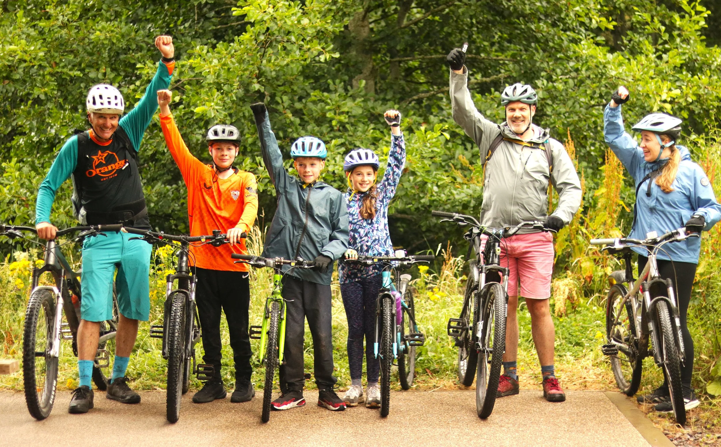 A group of six people, including children and adults, standing with their bikes on a paved pathway in a forested area. They are wearing helmets and outdoor attire and are raising their fists in celebration.