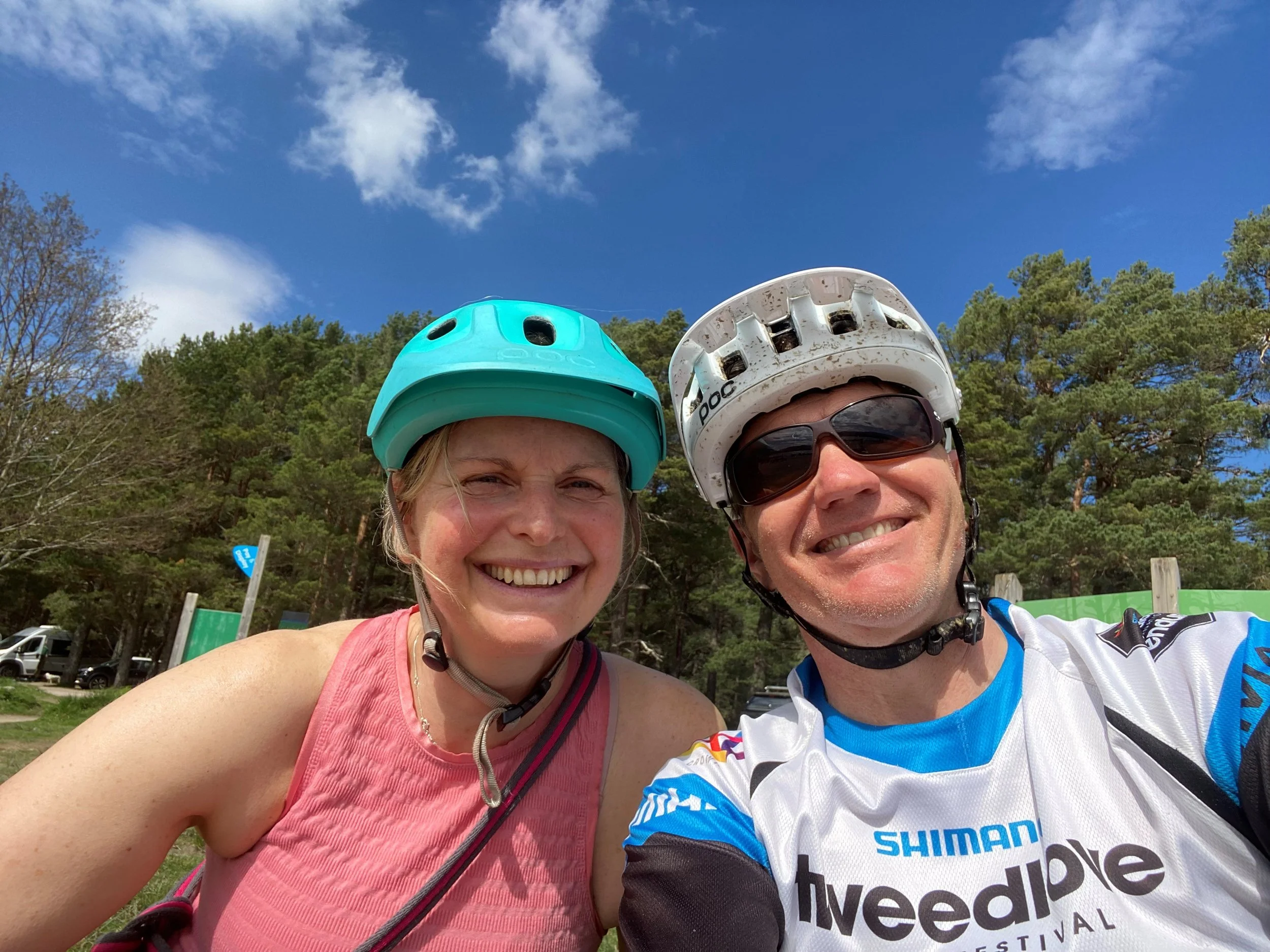 A smiling woman and man wearing helmets and sunglasses taking a selfie outdoors with a background of trees and a blue sky.