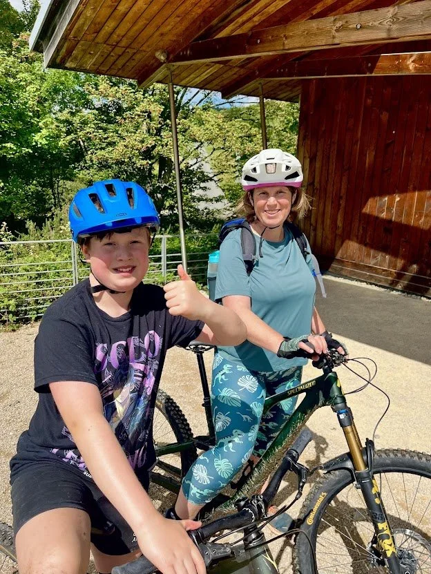 A boy and a woman smiling with helmets on, sitting on a mountain bike outdoors on a sunny day, near a wooden structure and green trees.