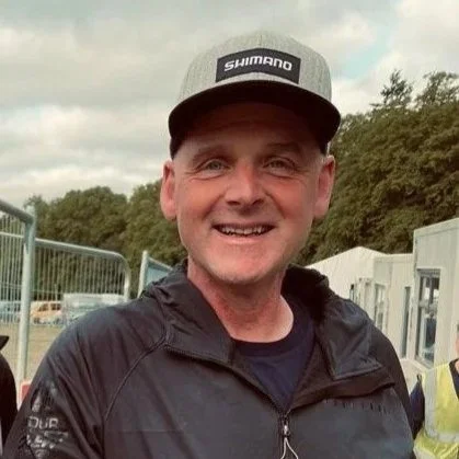 A smiling man wearing a gray Shimano cap and black jacket outdoors.