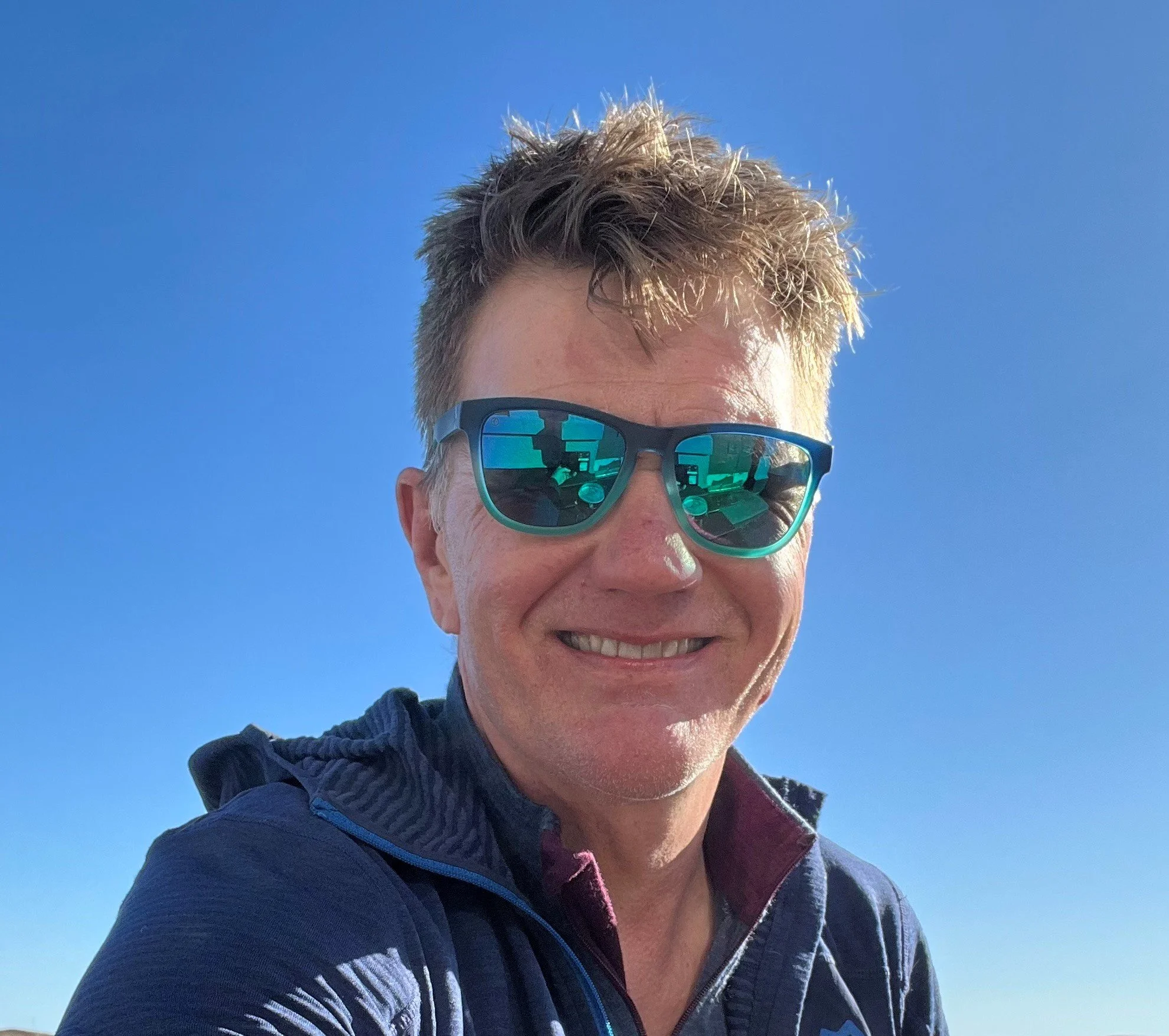 A smiling man with short, spiked hair wearing blue sunglasses and a blue jacket against a clear blue sky.