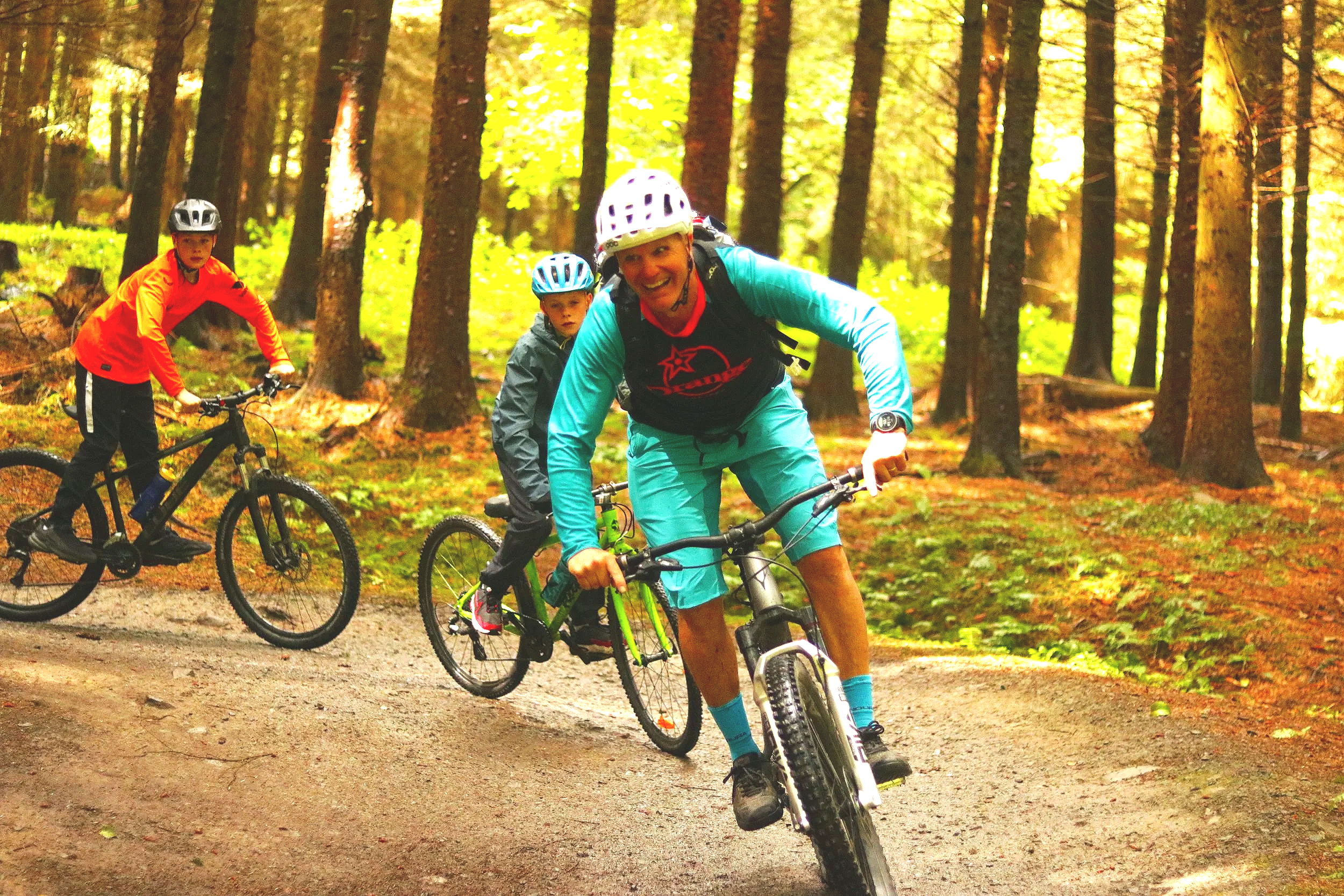 A smiling adult man wearing a white helmet, teal and black outfit, and a black backpack, riding a mountain bike on a trail through a forest with tall trees and green foliage, with two young children nearby also biking and wearing helmets.