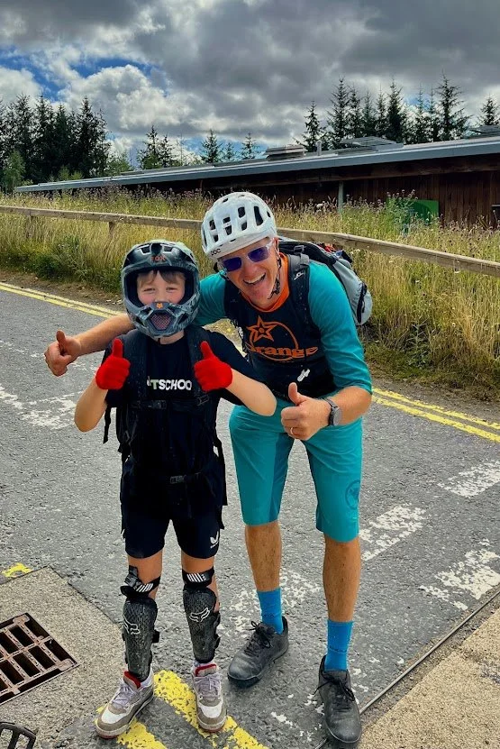 A smiling child and a guide outdoors on a paved road, both wearing helmets and protective gear, giving thumbs up.