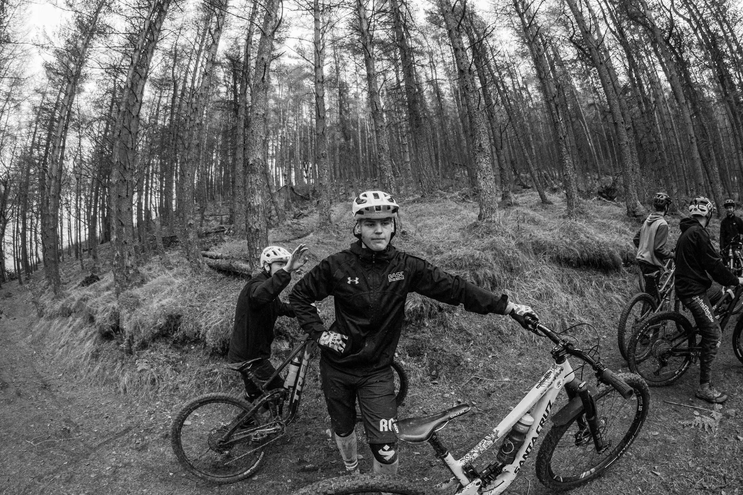 Group of cyclists in helmets and sportswear in a wooded forest, some standing with bikes, one adjusting his bike, in black and white.