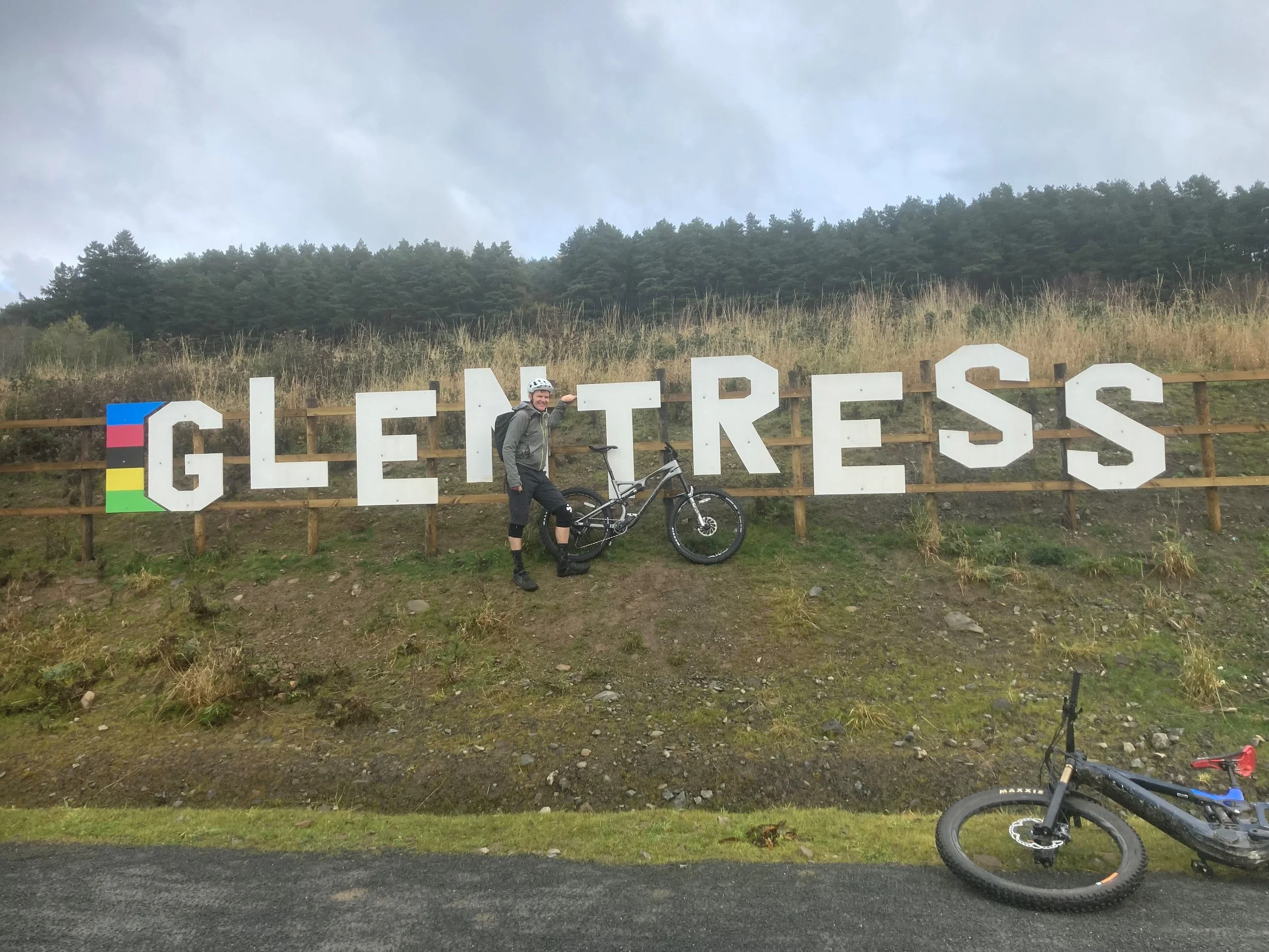 Bike trail sign reading 'Gletress' with a person in cycling gear pointing at it, and a bicycle standing on the ground nearby, with a second bike lying on the road.