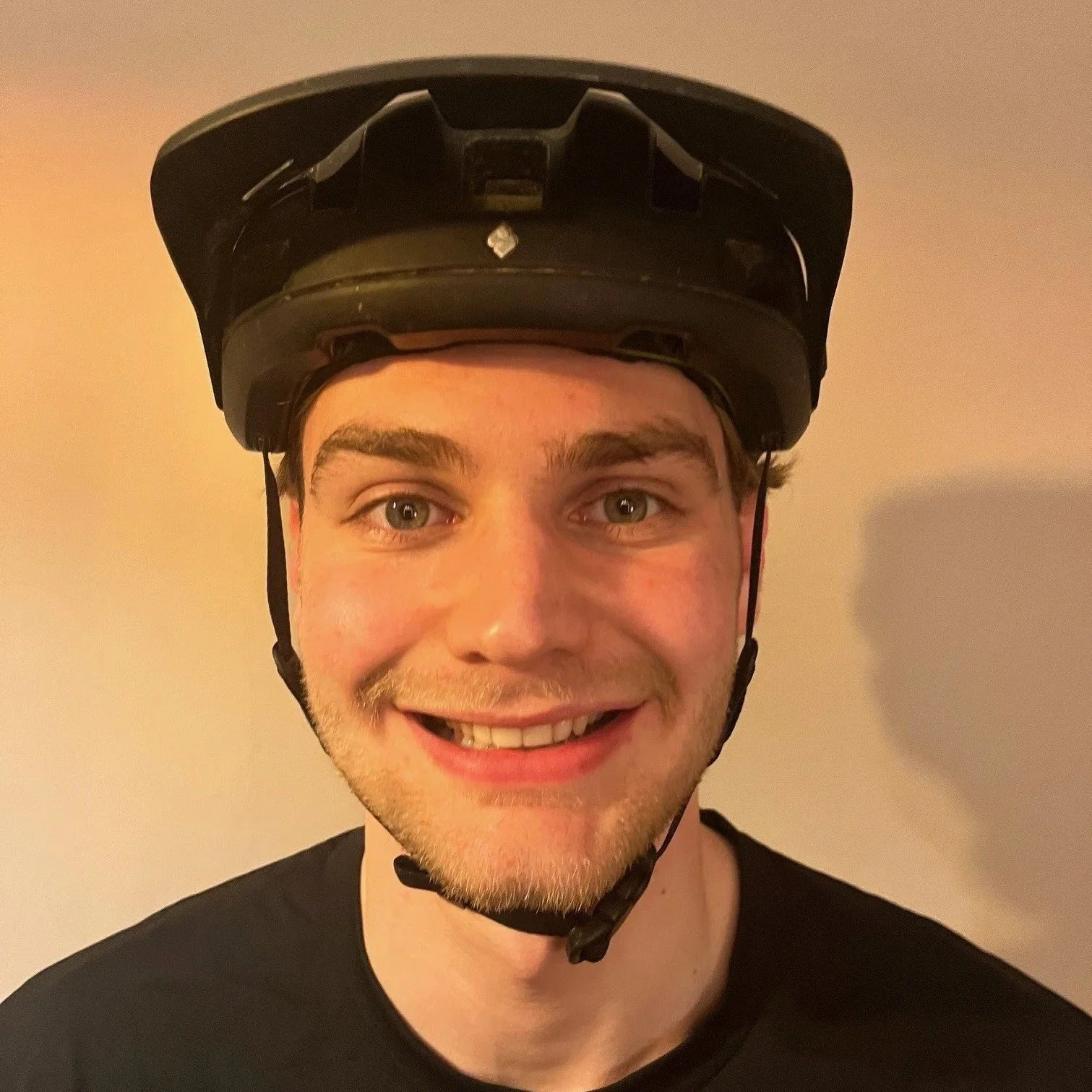 A young man smiling with snow on his face, wearing a black bicycle helmet, standing indoors against a plain wall.