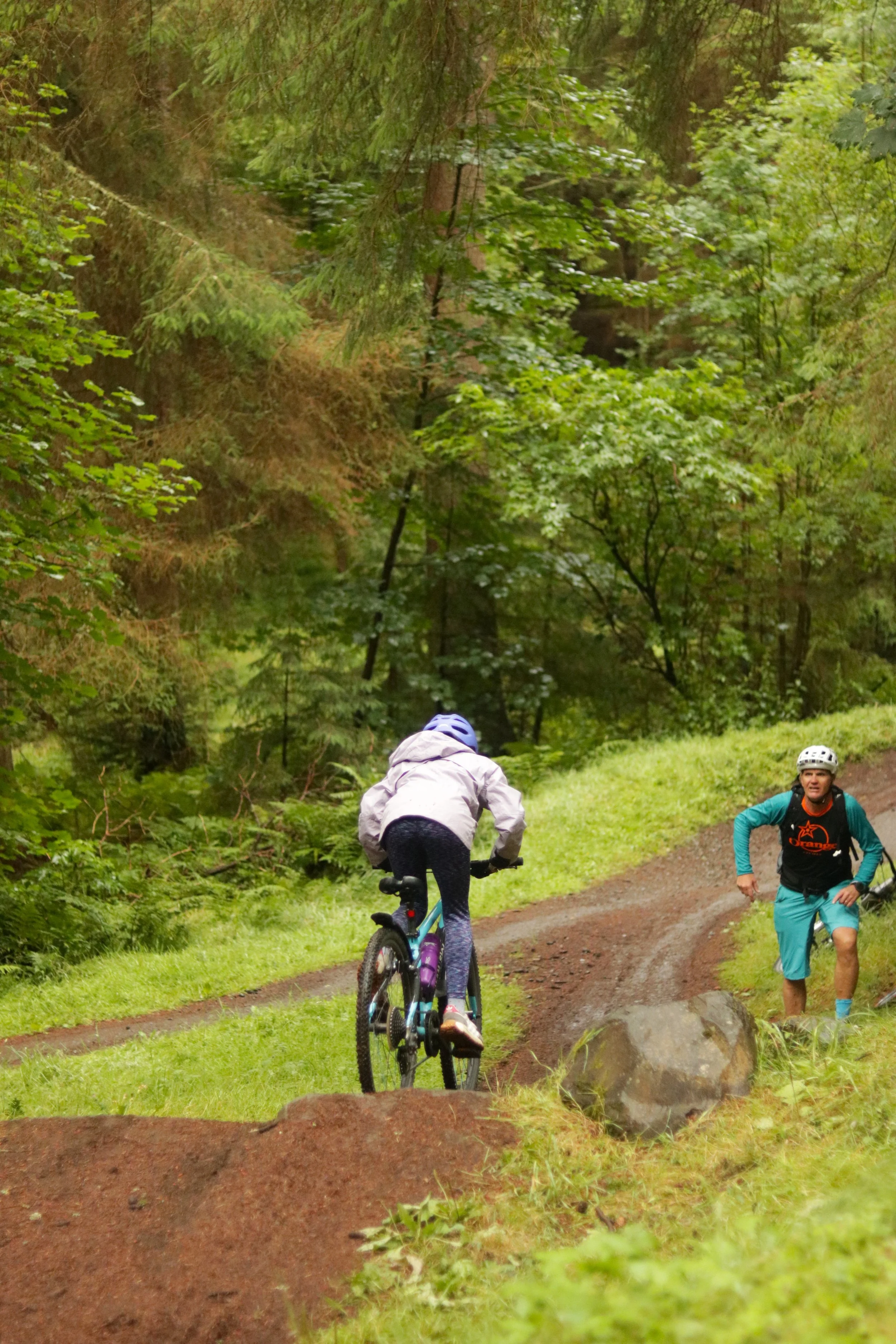A person riding a bicycle along a dirt trail in a forest, with a man running nearby on the trail, surrounded by lush green trees and foliage.