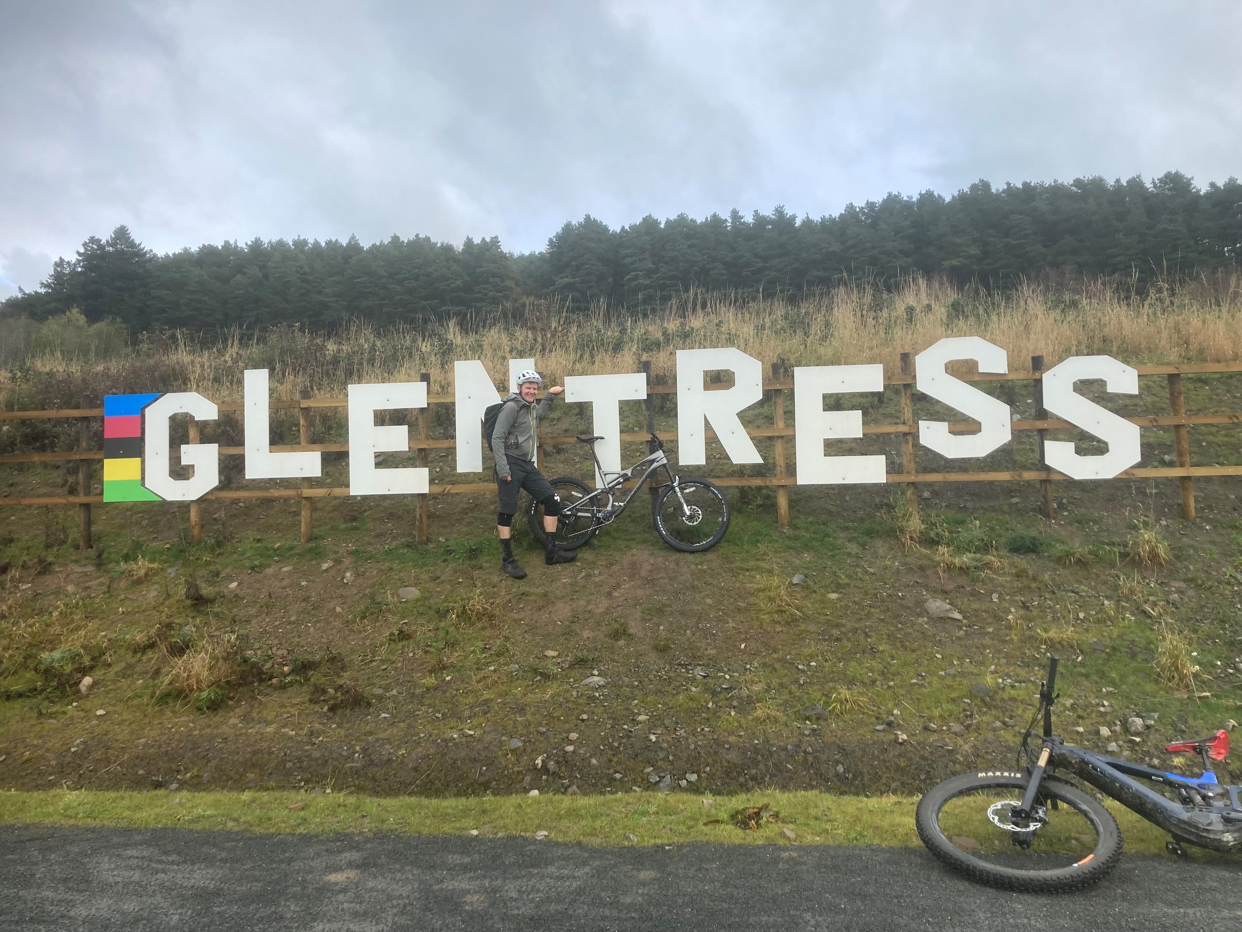 Person in cycling gear with a helmet leaning on a mountain bike next to a large sign that reads 'GLETTERS' with colored stripes on the left, in front of a grassy hill and trees.