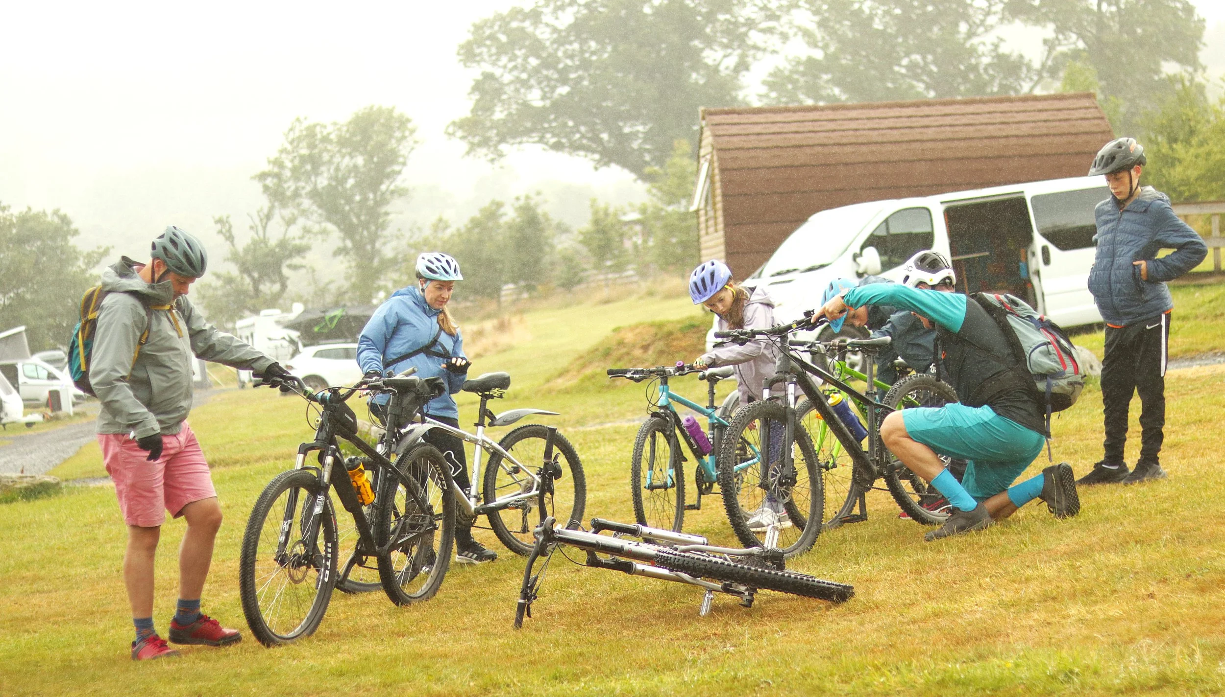 Group of people with bicycles outdoors near a small building and a van, some wearing helmets, in a foggy grassy area.
